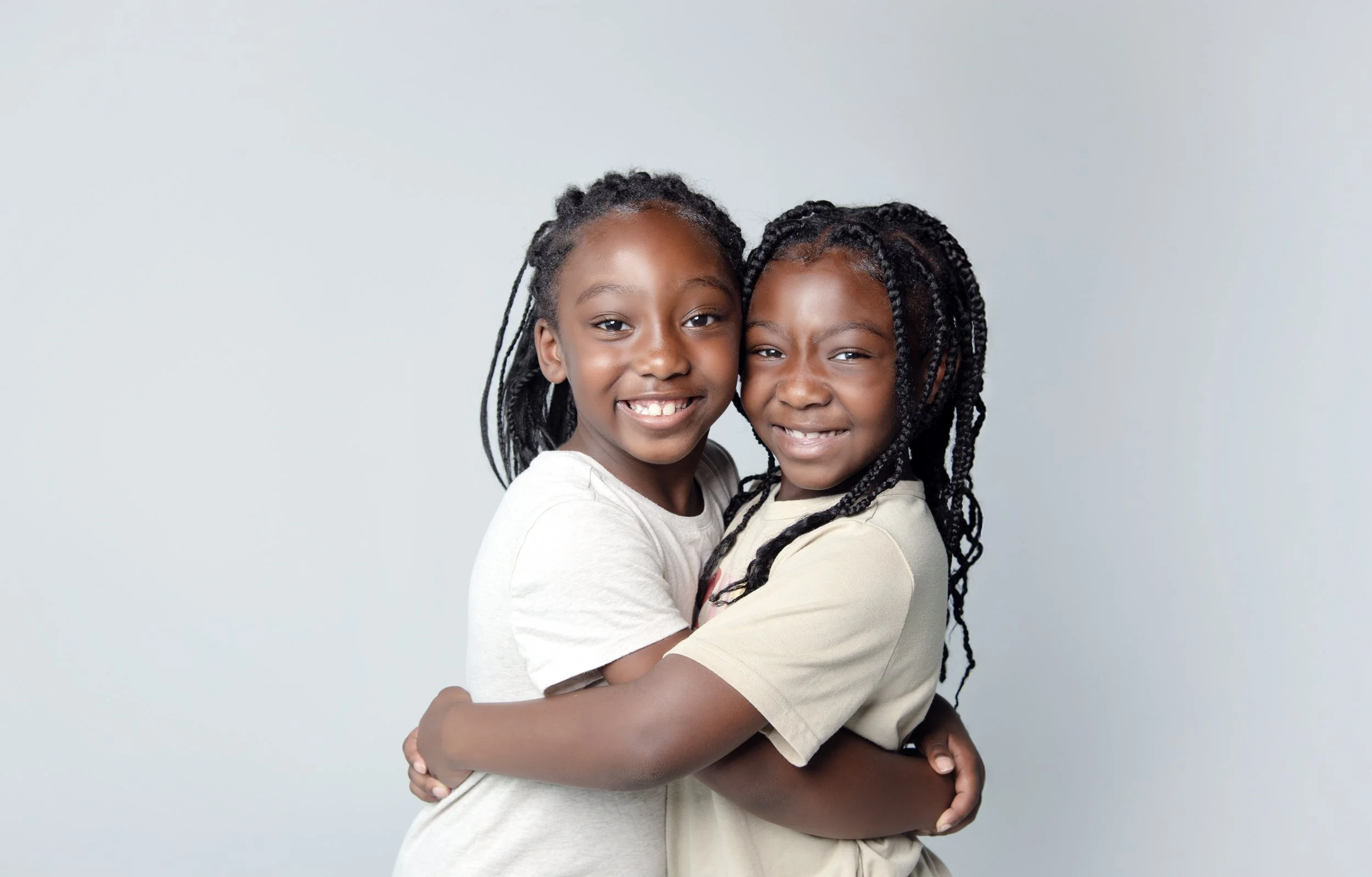 Two young girls with braided hair hugging and smiling at the camera against a plain light gray background.