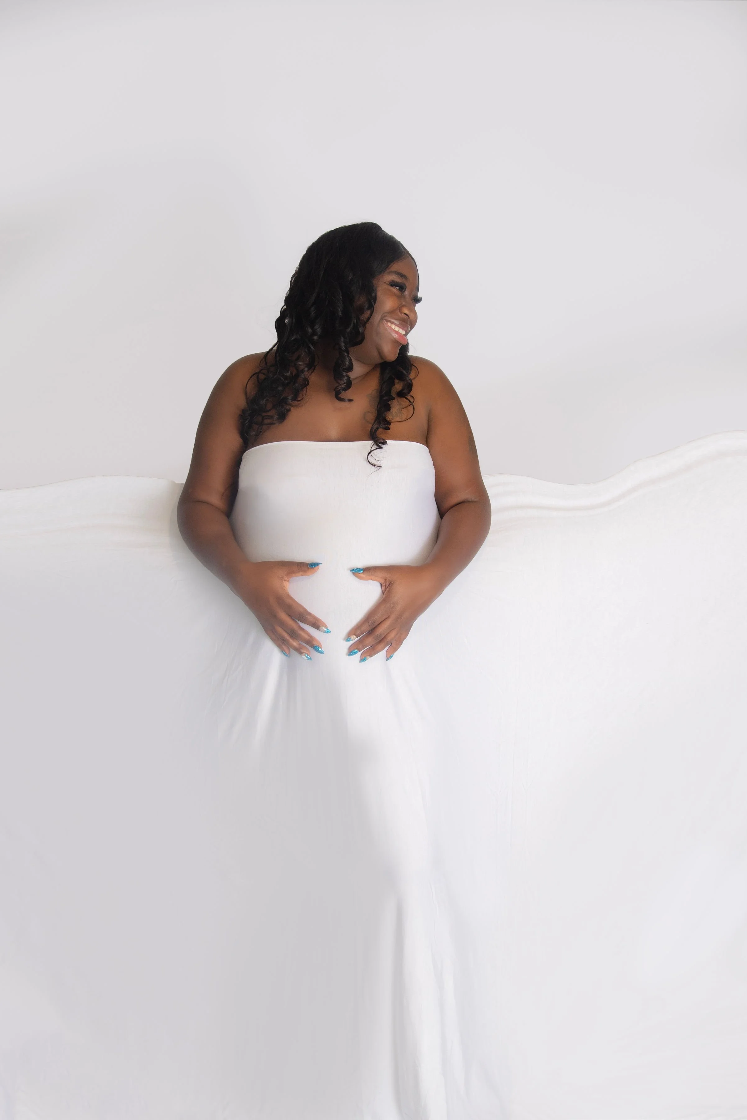 A smiling woman with curly hair wearing a white strapless dress, standing against a plain white background, with her hands gently placed on her belly.