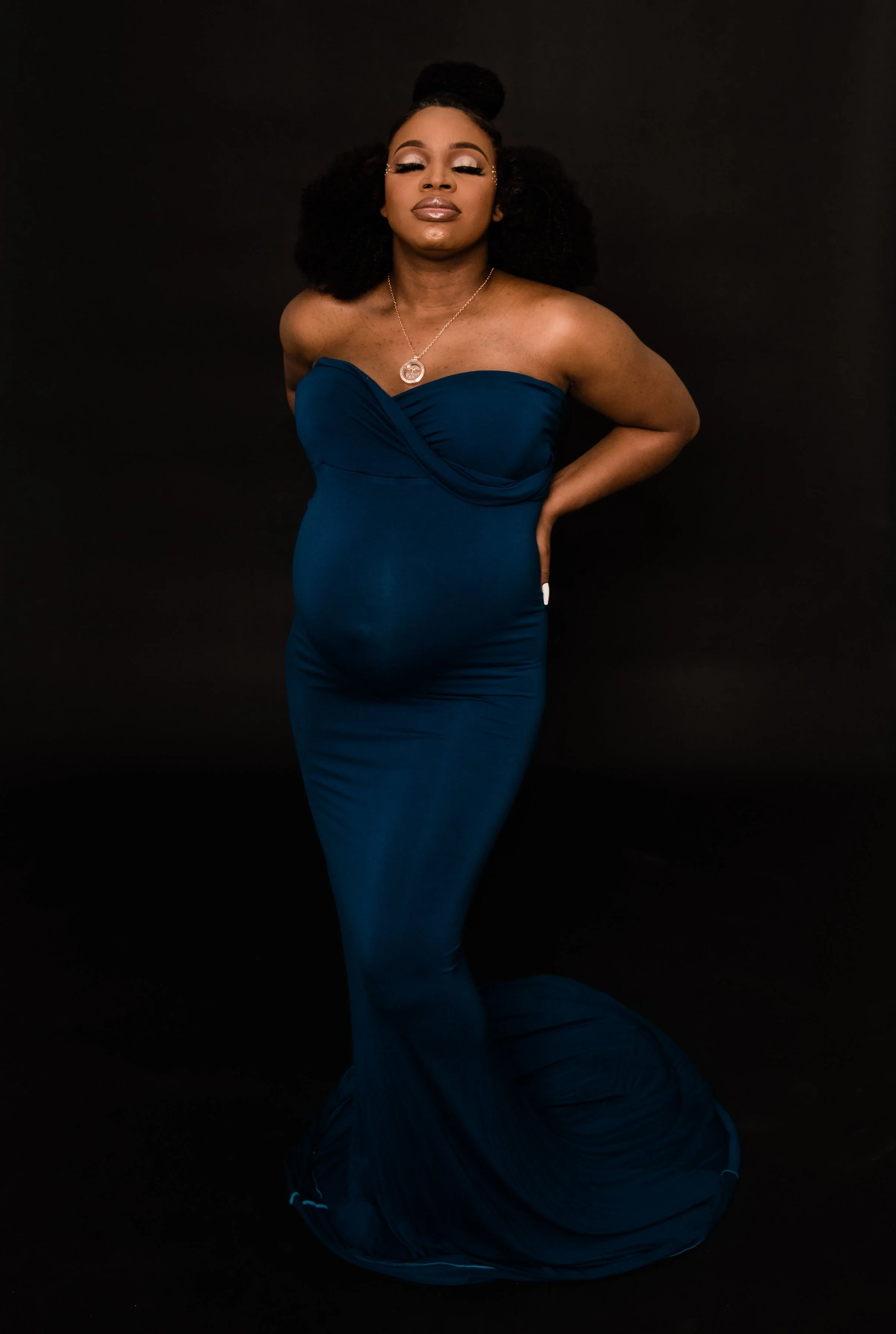 A woman in a strapless blue gown standing against a black background, with one hand on her hip and eyes closed.