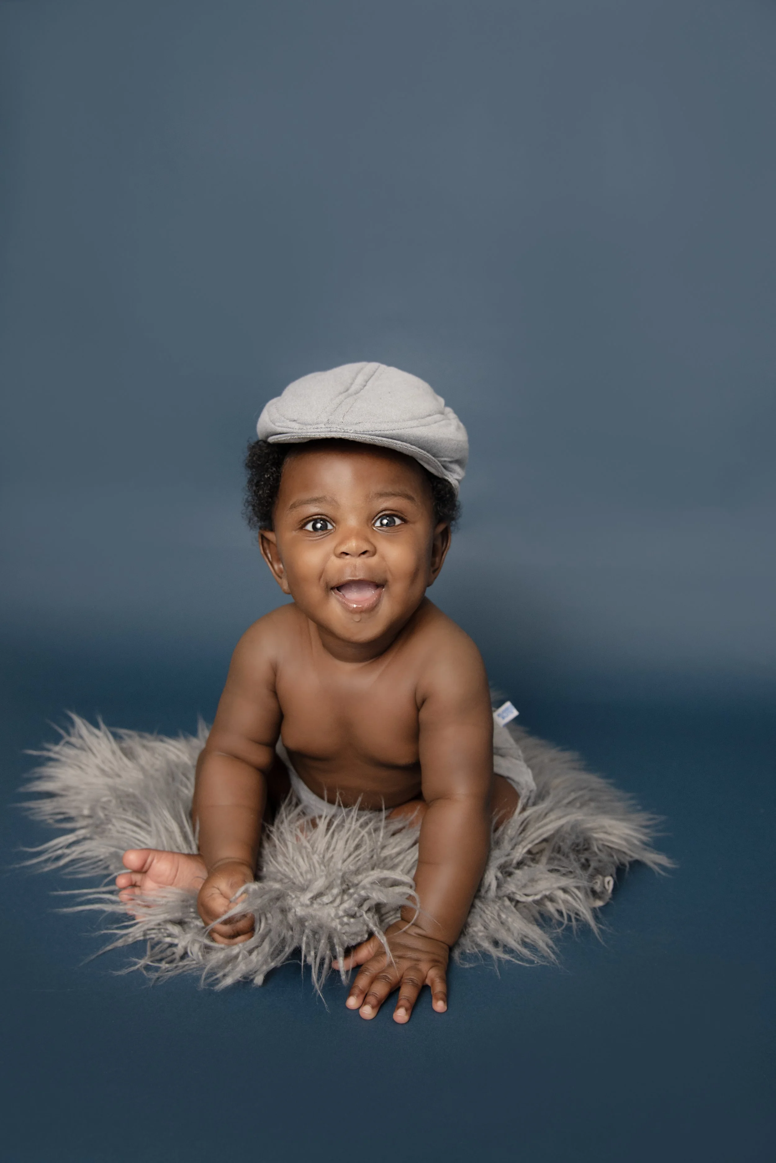A smiling toddler wearing a gray cap, sitting on a furry gray blanket against a blue background.