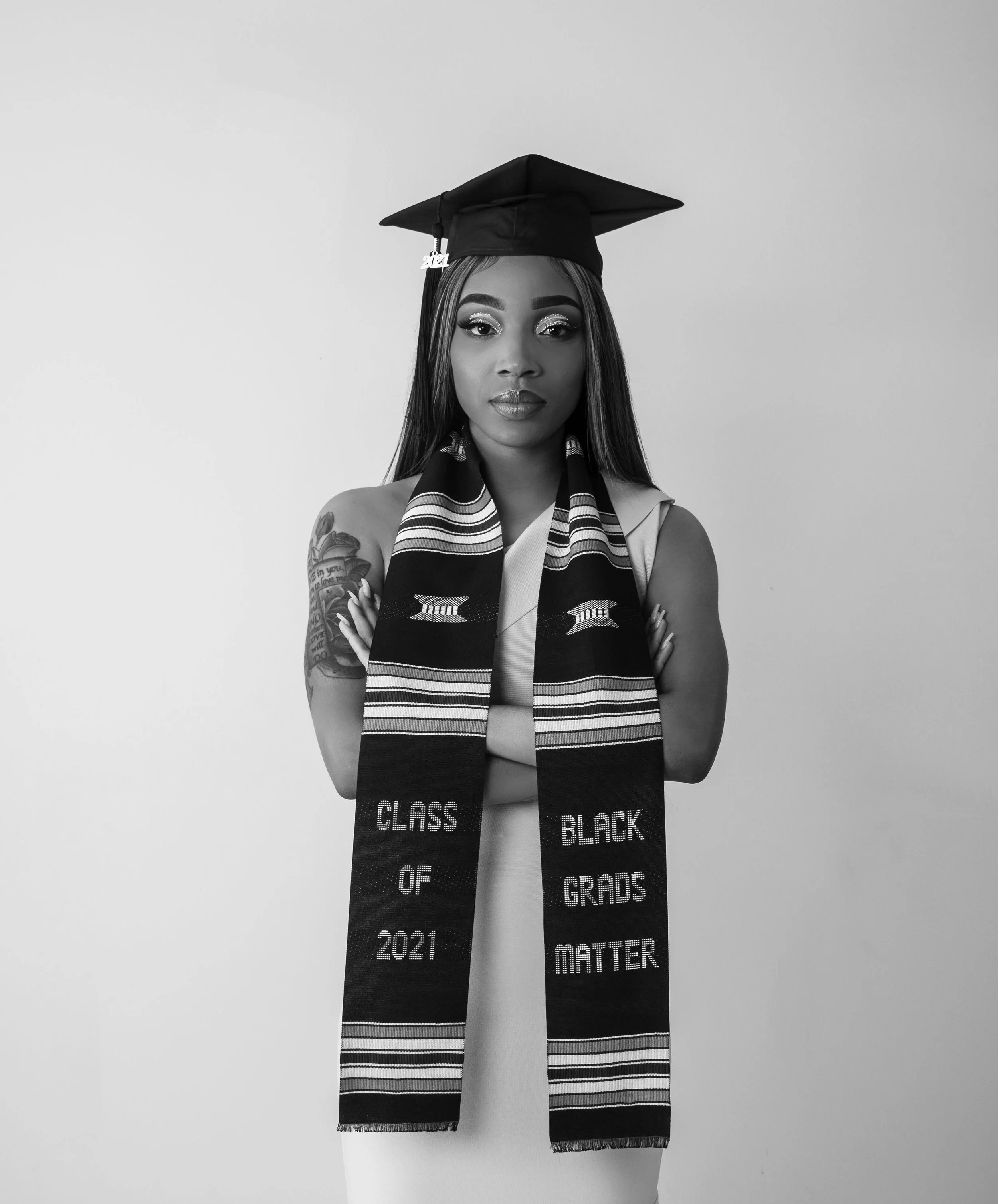 A Black woman in a graduation cap and stole, with arms crossed, standing against a plain background.