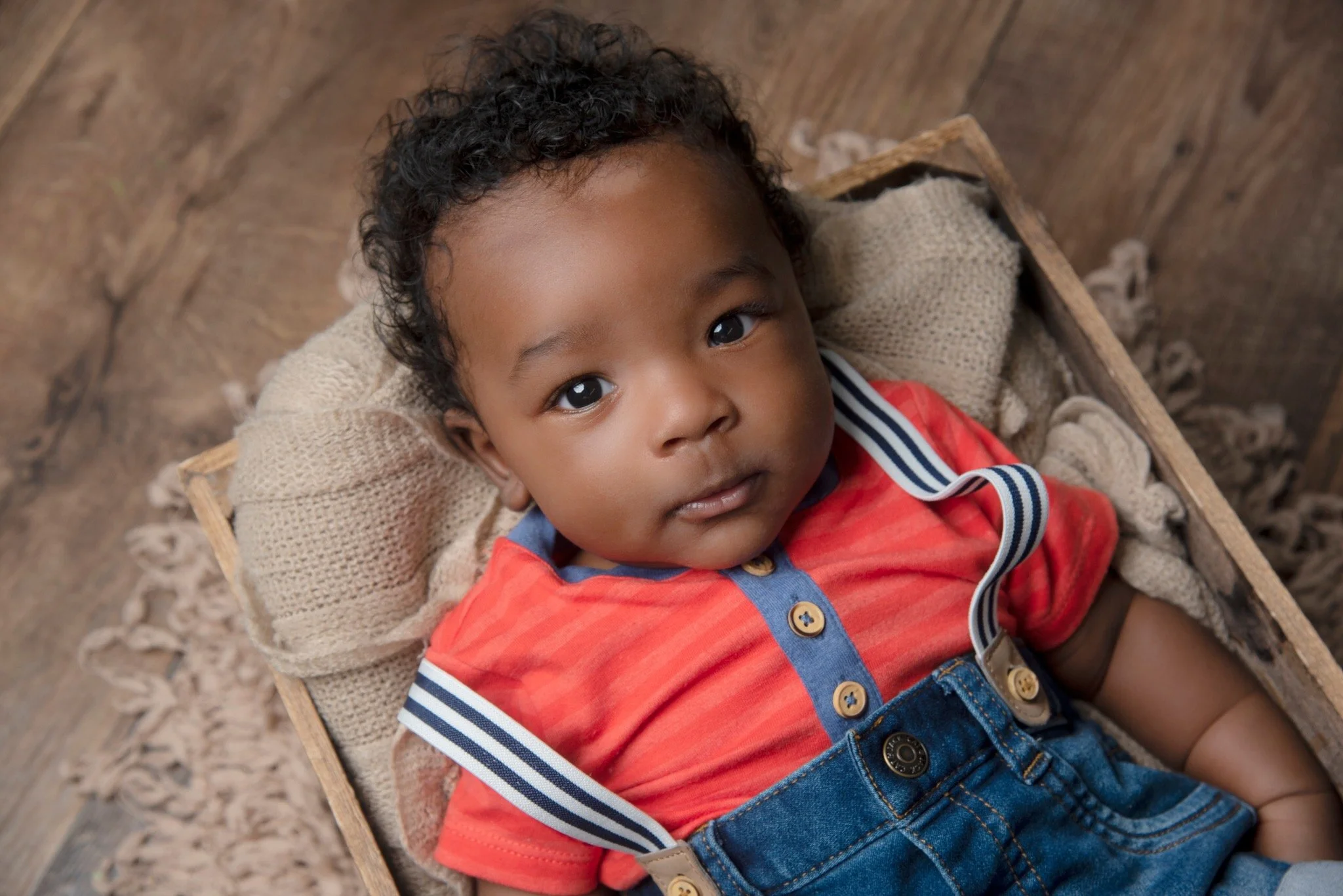 A young boy lying in a woven basket with a beige blanket, wearing a red and blue collared shirt and denim overalls, looking up.
