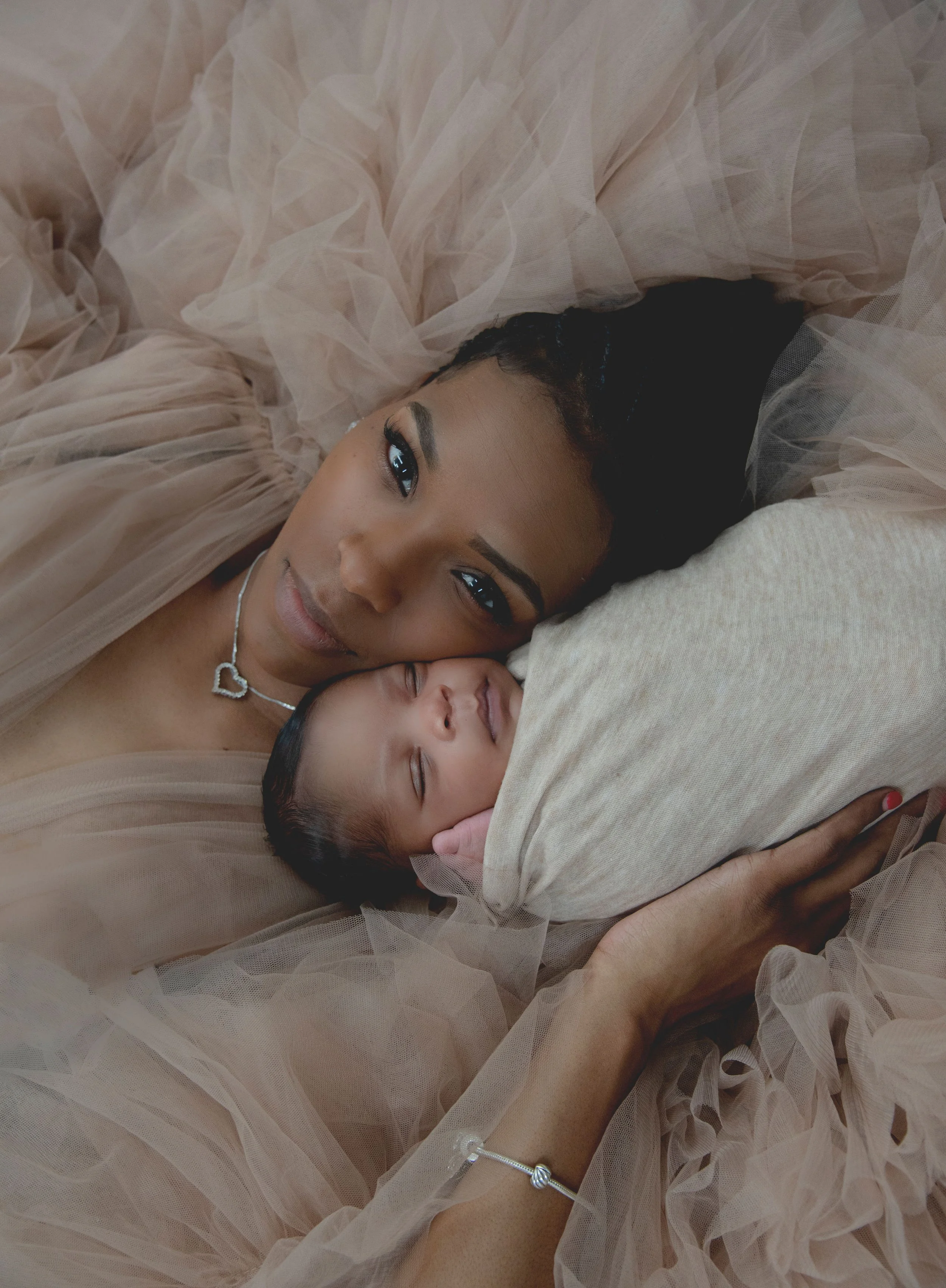 A woman and a sleeping baby lying on a cream pillow, with soft, beige tulle fabric surrounding them.