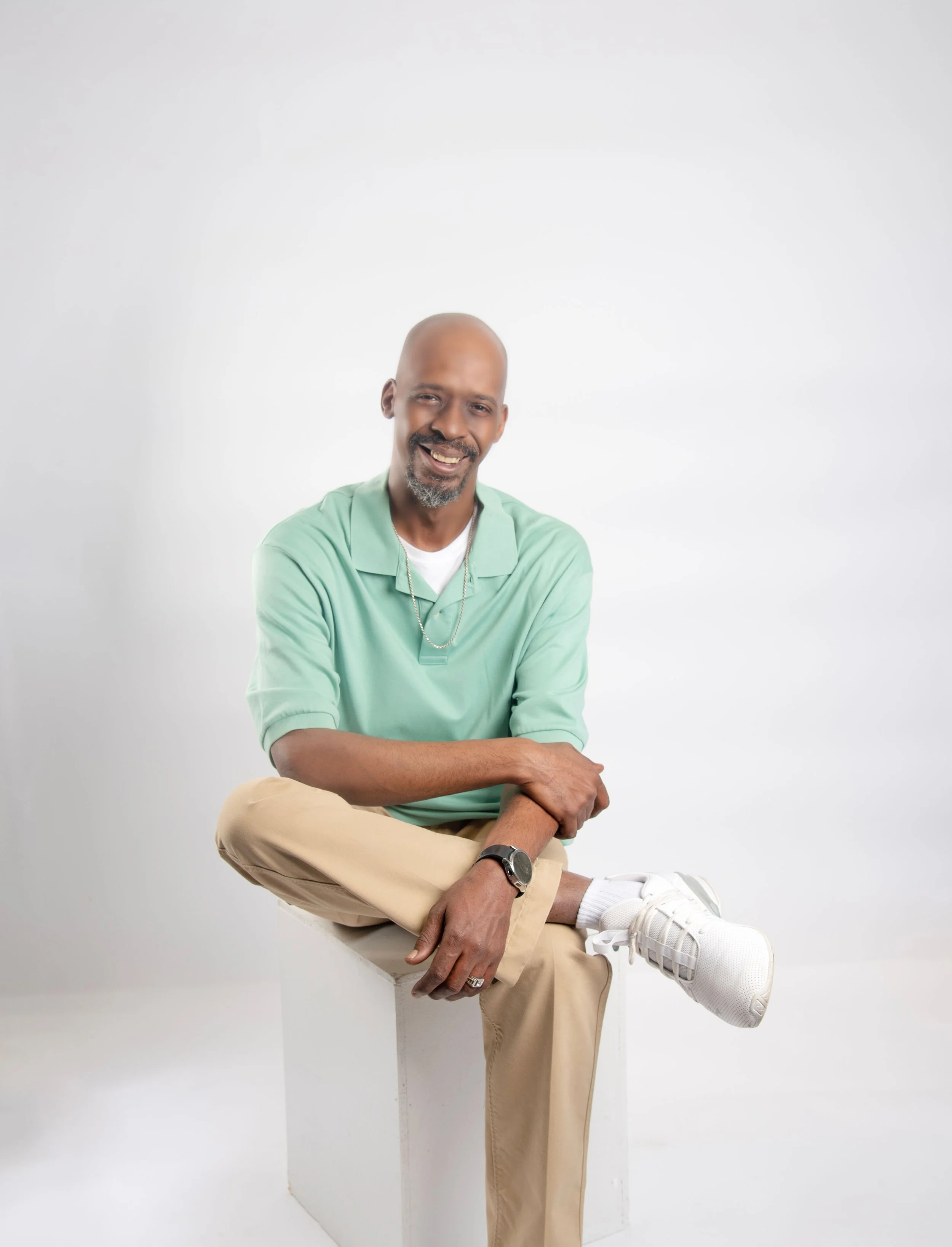Smiling man with bald head and goatee, wearing light green polo shirt, tan pants, white sneakers, and watch, sitting cross-legged on a white block against a plain white background.