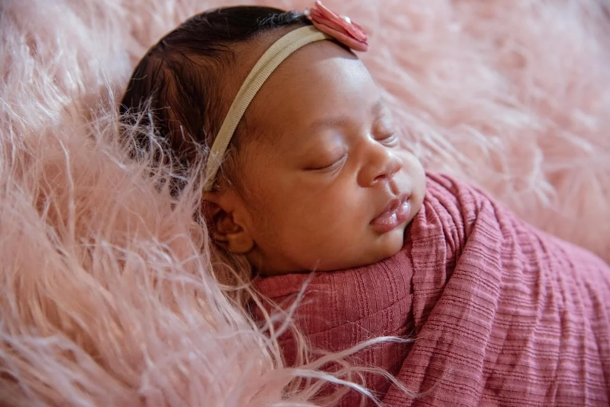 A sleeping newborn baby wrapped in a pink blanket, wearing a beige headband with a pink flower, lying on a pink fluffy surface.