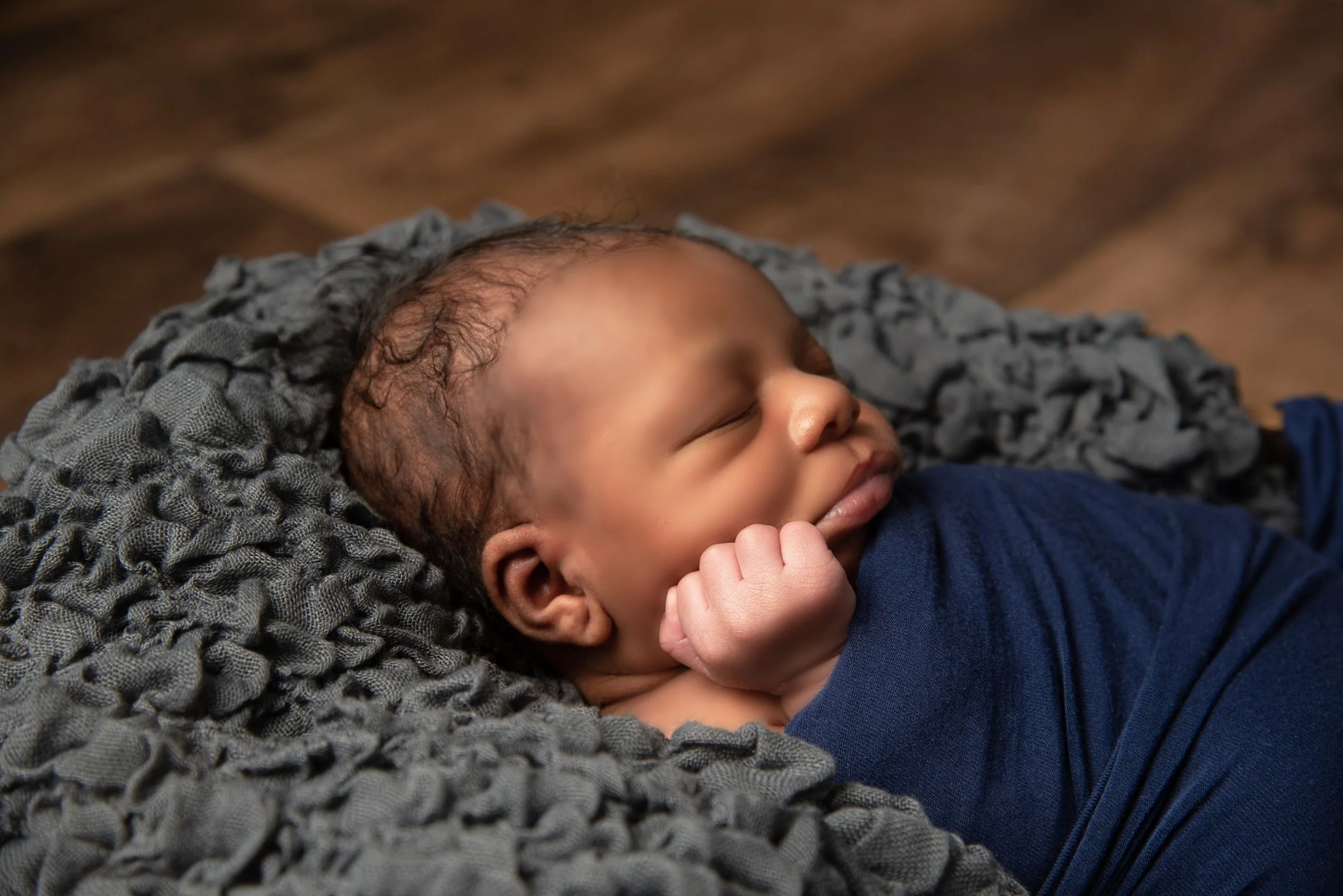 A sleeping baby with curly hair lying on a soft gray blanket, wearing a blue shirt.