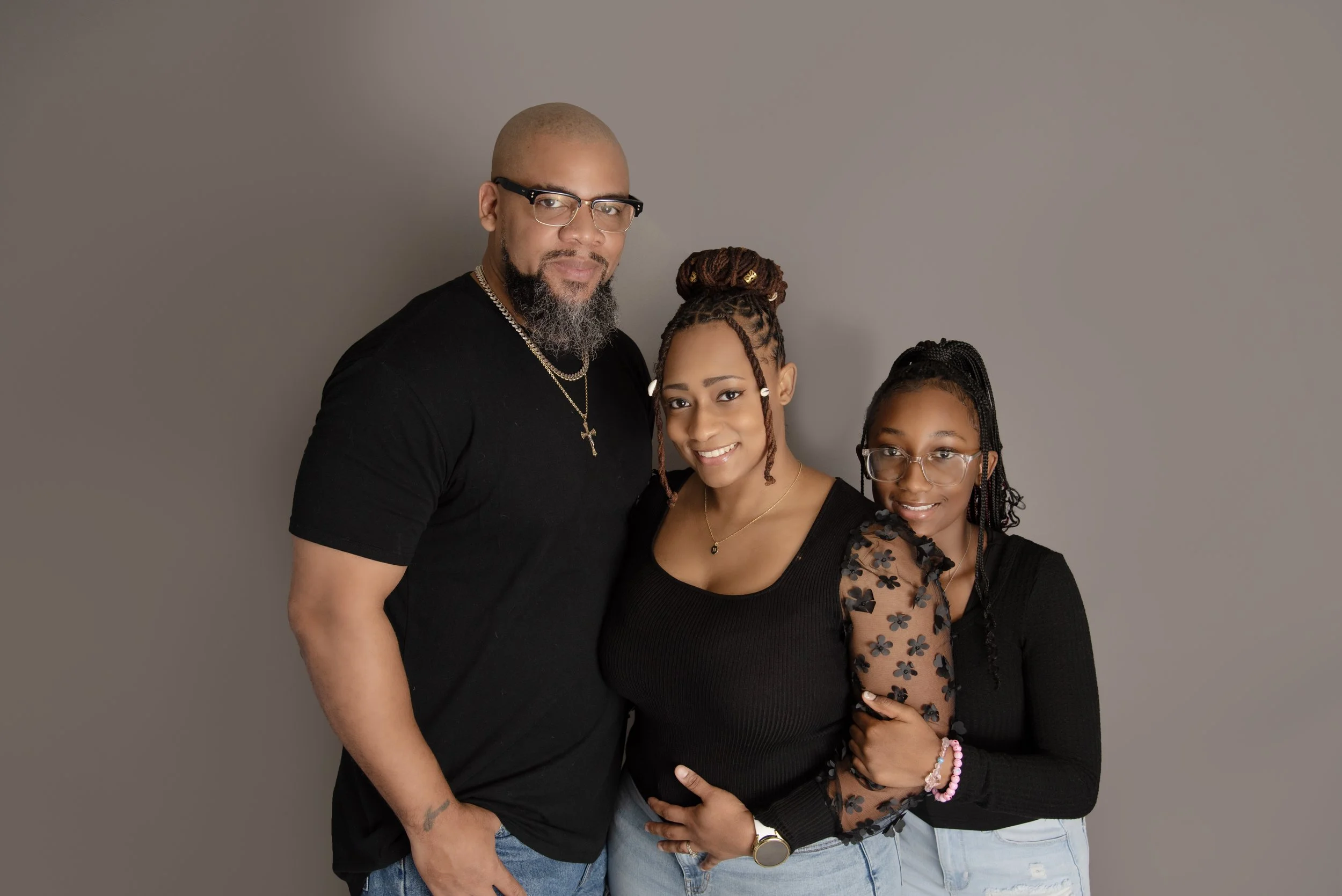 A man with glasses, a beard, and a shaved head standing next to a woman with dreadlocks in a bun, and a young girl with glasses, all smiling and standing close together against a gray background.