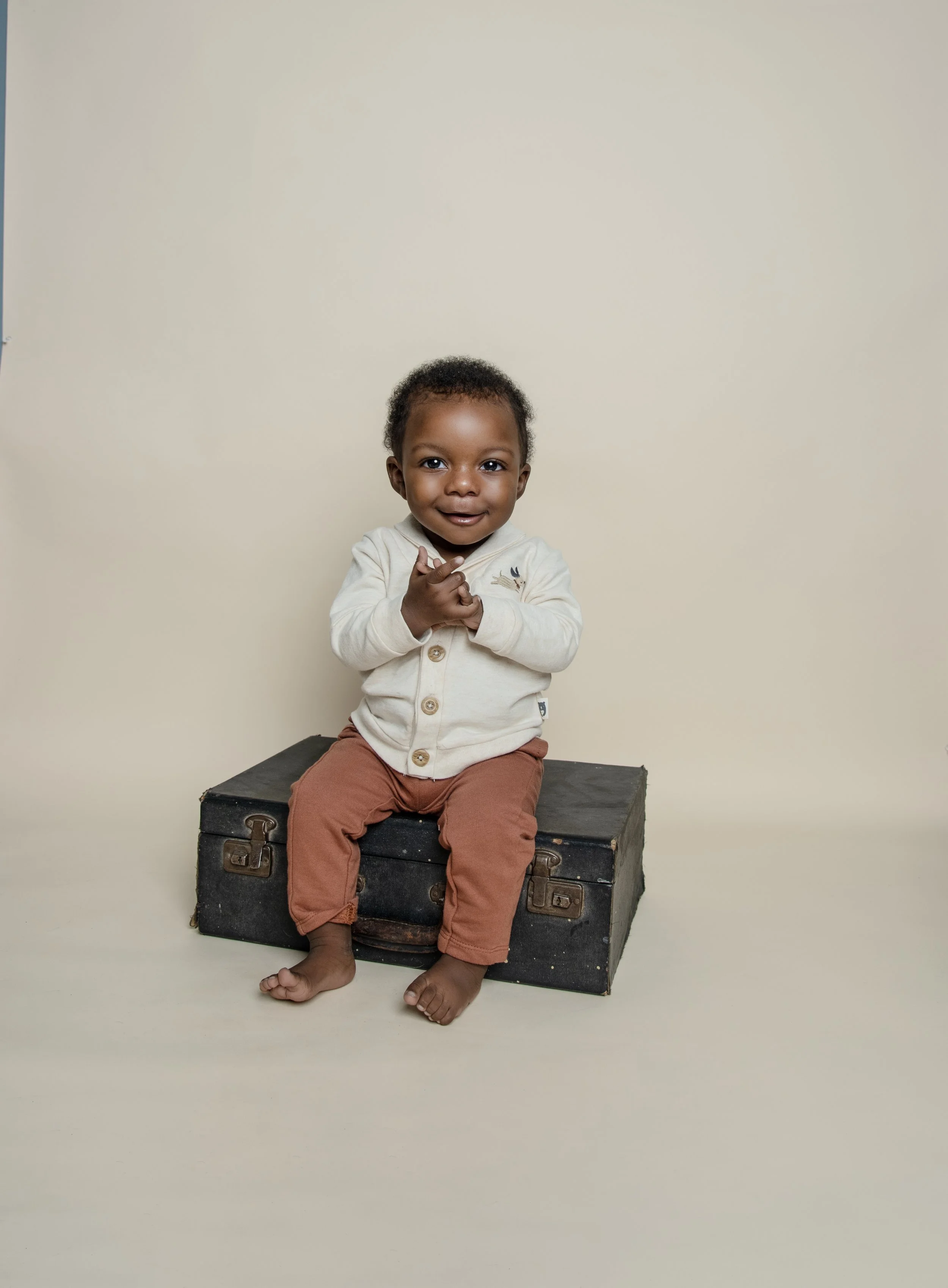A young child with dark curly hair, sitting barefoot on an old black suitcase, dressed in a cream-colored cardigan and rust-colored pants, holding hands together and smiling at the camera against a plain cream background.