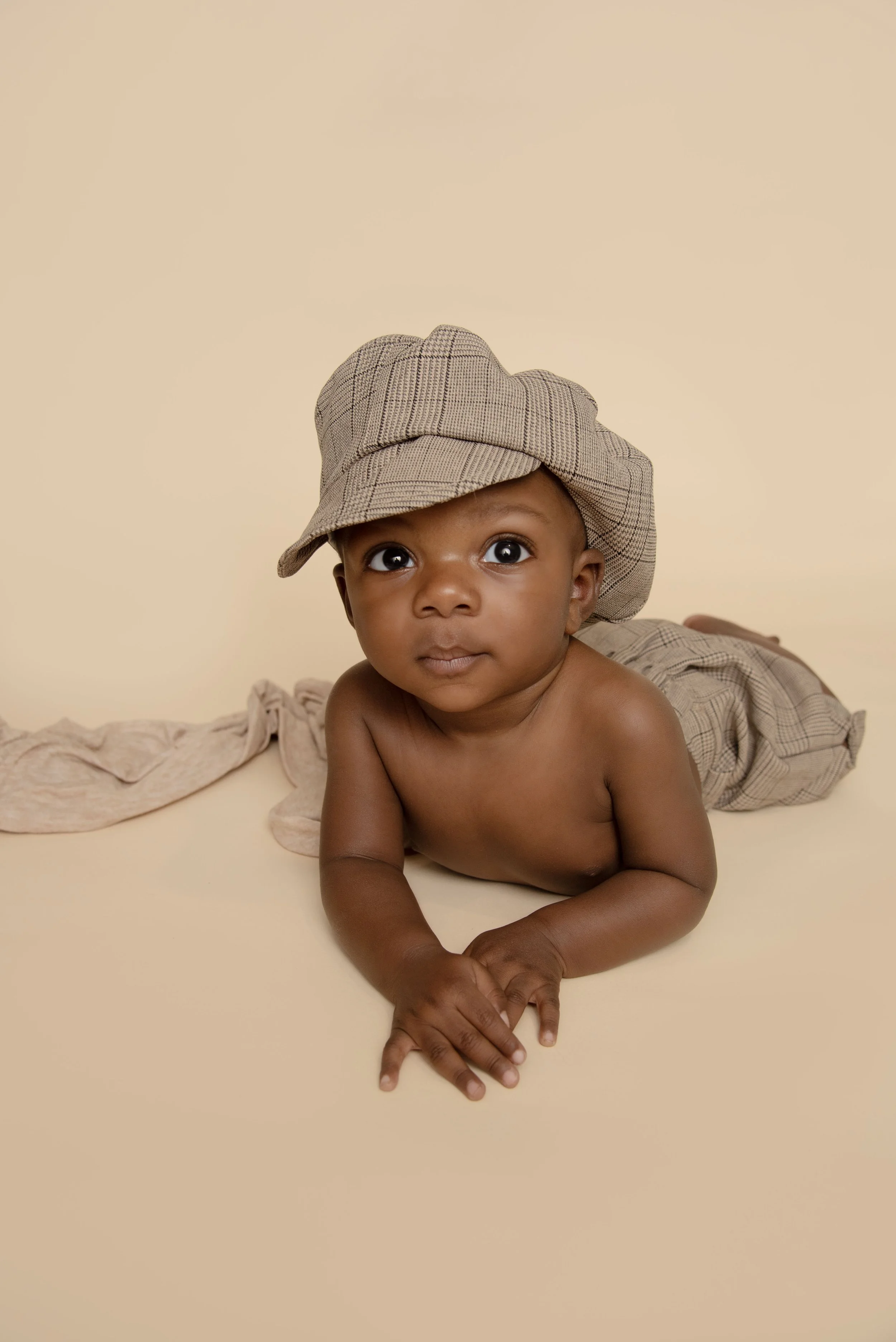 A young child with large eyes lying on their stomach against a soft beige background, wearing a plaid hat and matching pants, holding their hands together.