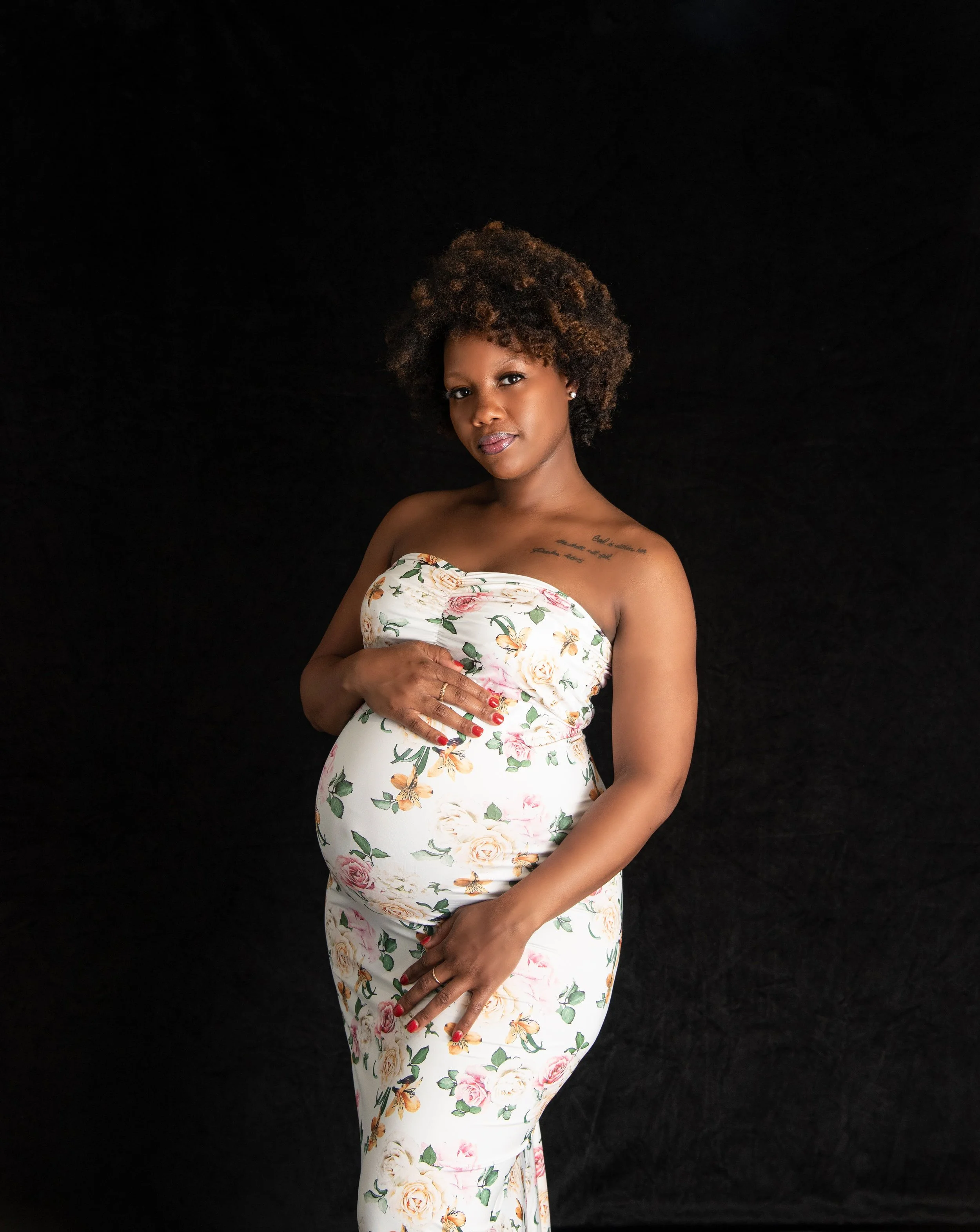Pregnant woman in a floral strapless dress standing against a black backdrop