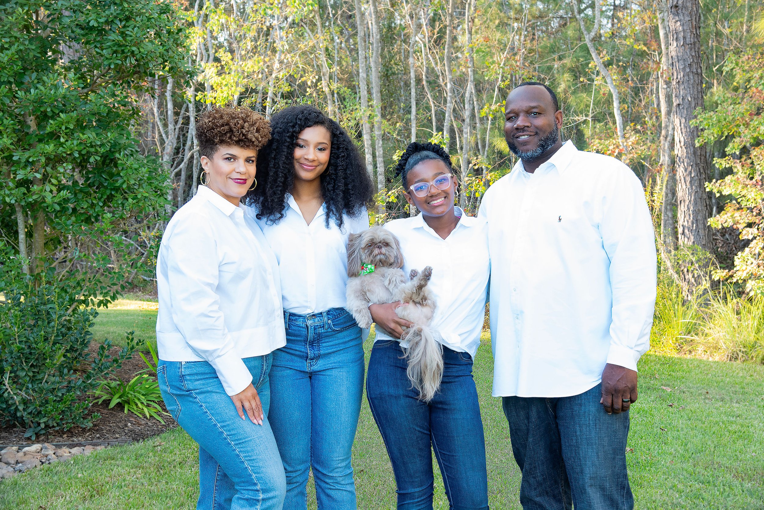 A family of five with a small dog standing outdoors in a grassy area with trees in the background. The family members are smiling and dressed in white shirts and blue jeans.