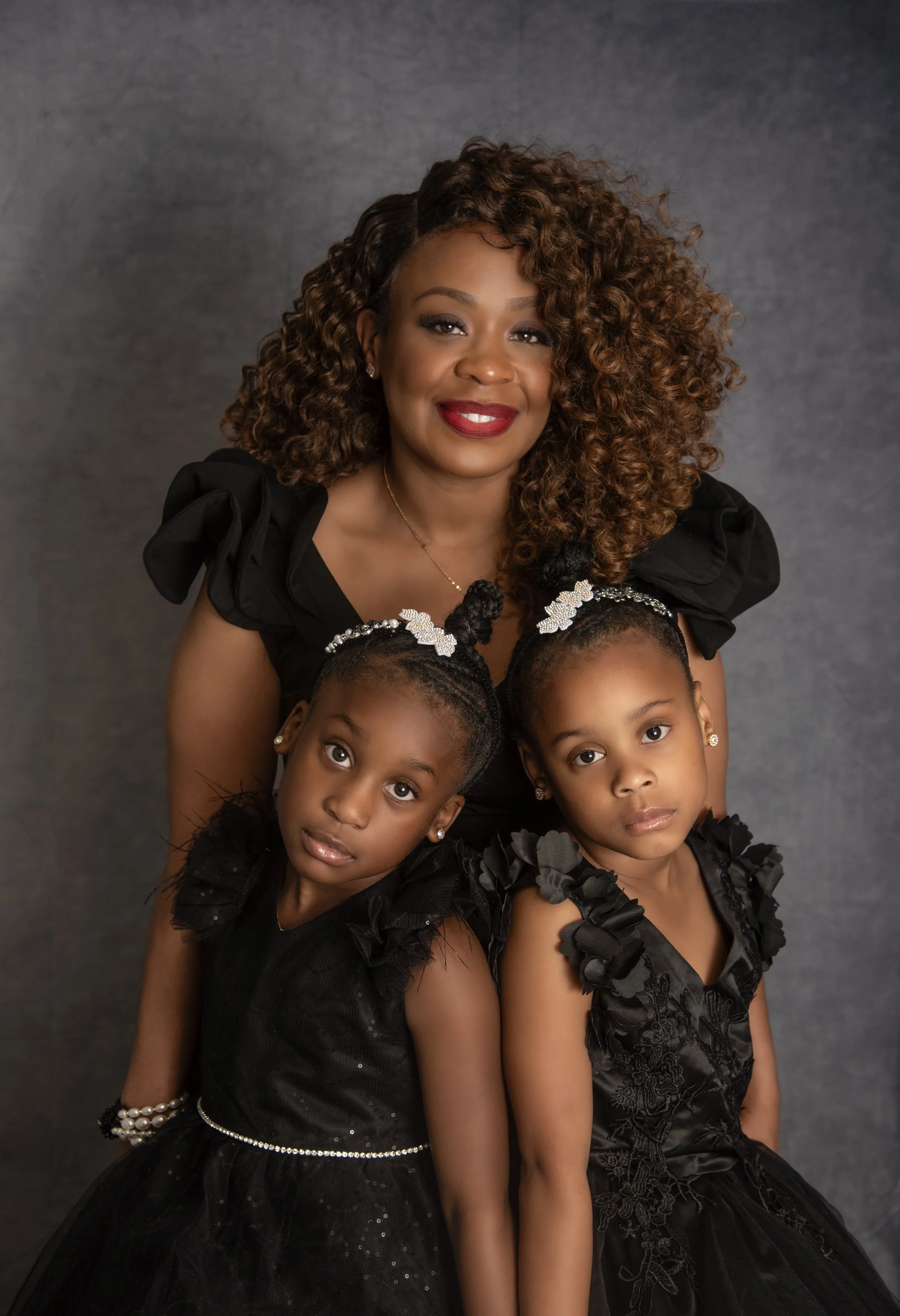 A woman with curly hair smiling in a black dress, with two young girls also in black dresses and matching hair accessories, posing against a gray background.