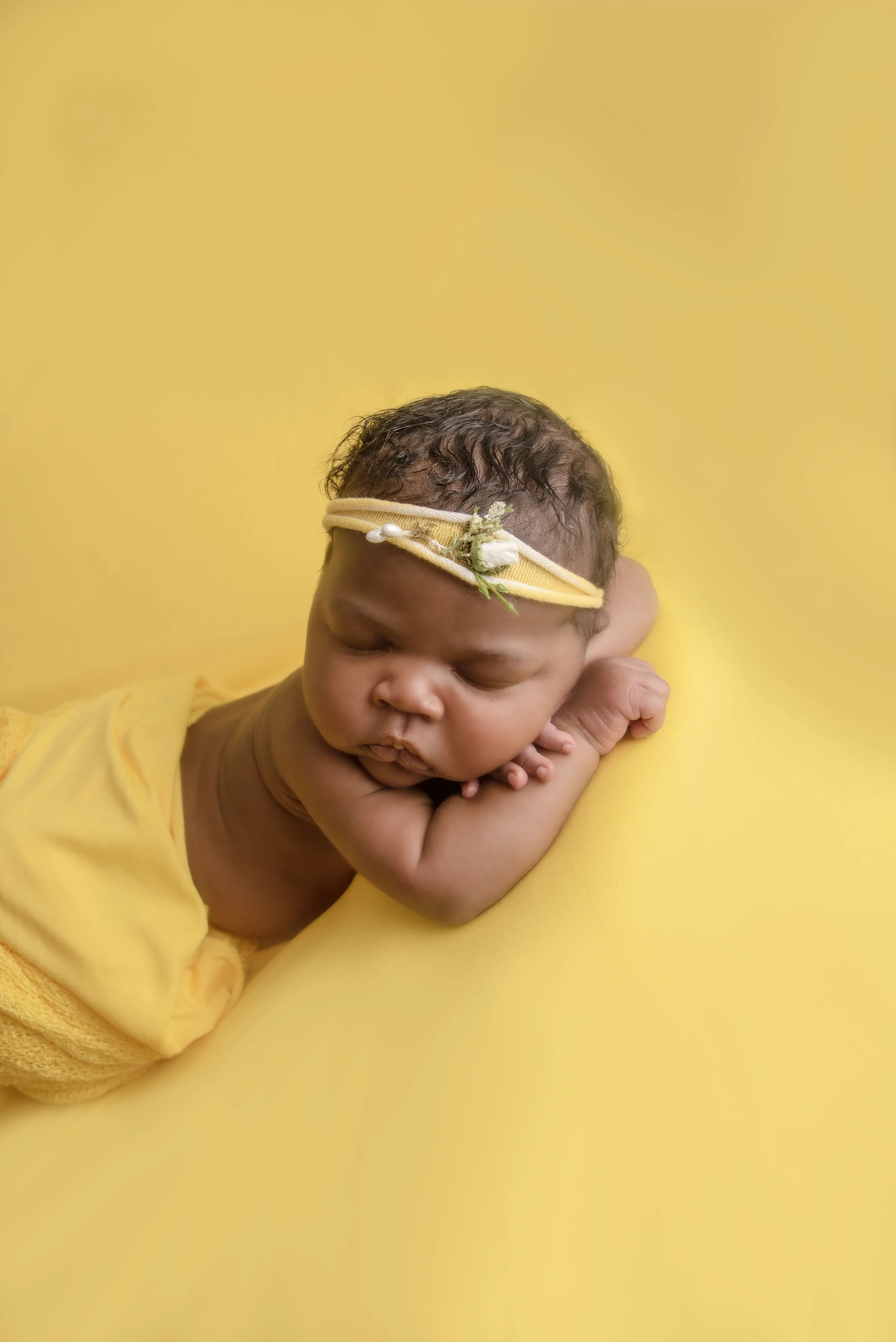A sleeping baby lying on a yellow surface, with head resting on arms, wearing a yellow headband with a small decorative flower, and wrapped in a yellow cloth.