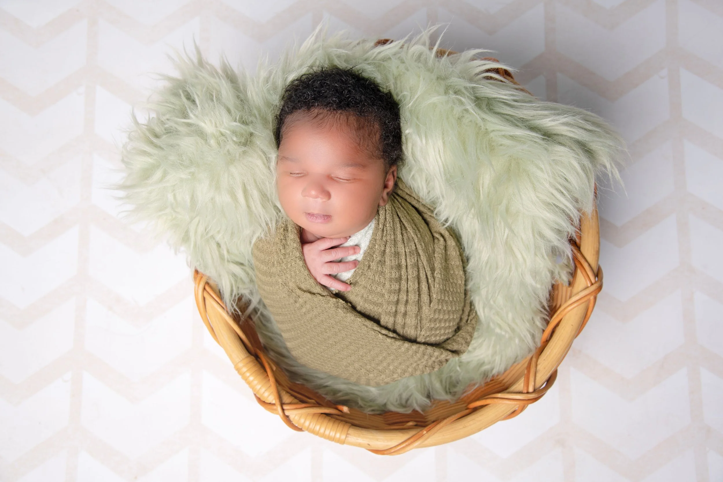 Close-up of a sleeping newborn baby in a wicker basket with a green furry blanket lining.