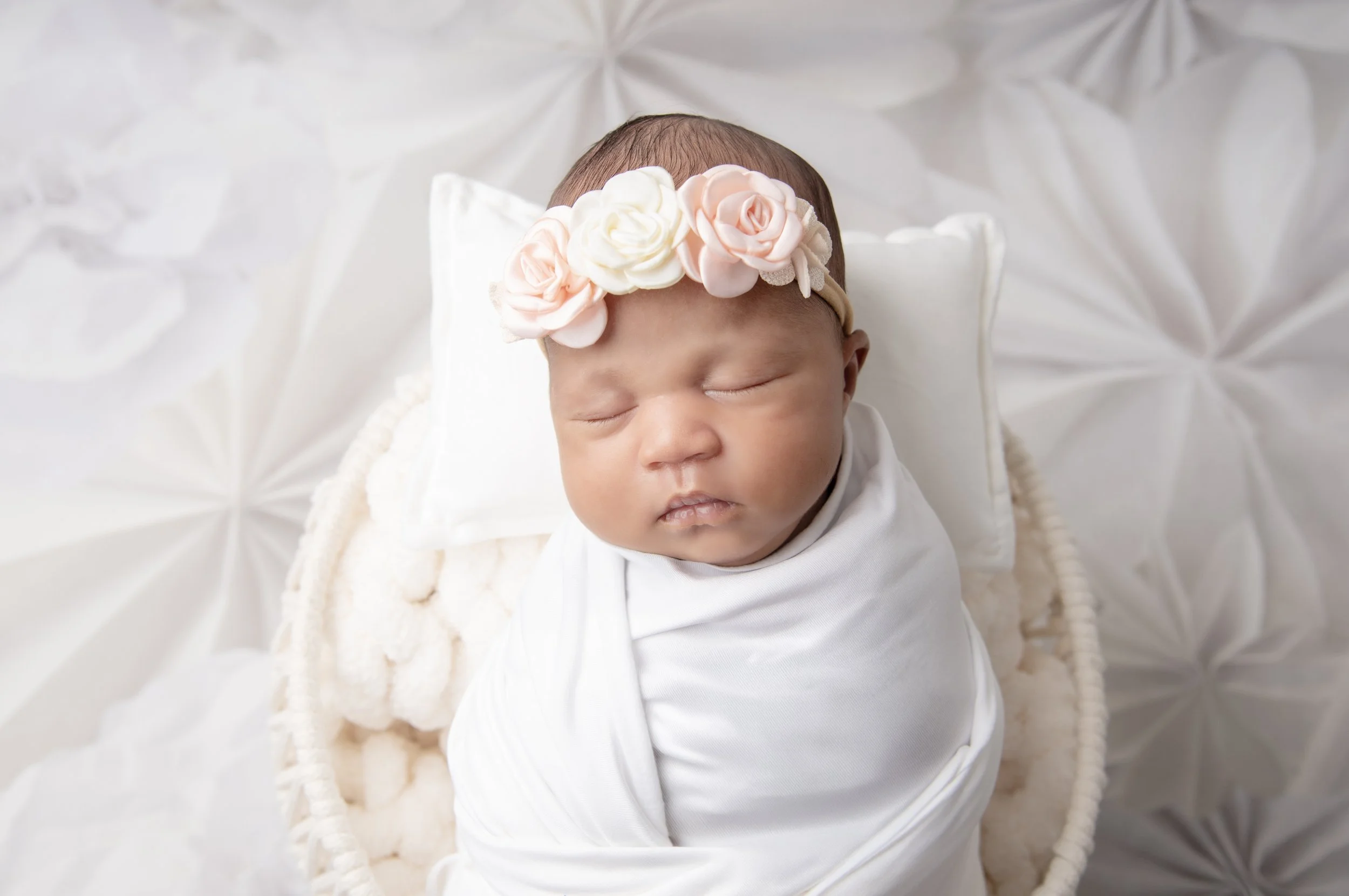 A sleeping newborn baby wrapped in a white blanket, wearing a pink and white flower headband, lying on a white pillow in a wicker basket with plush blanket, against a textured white background.
