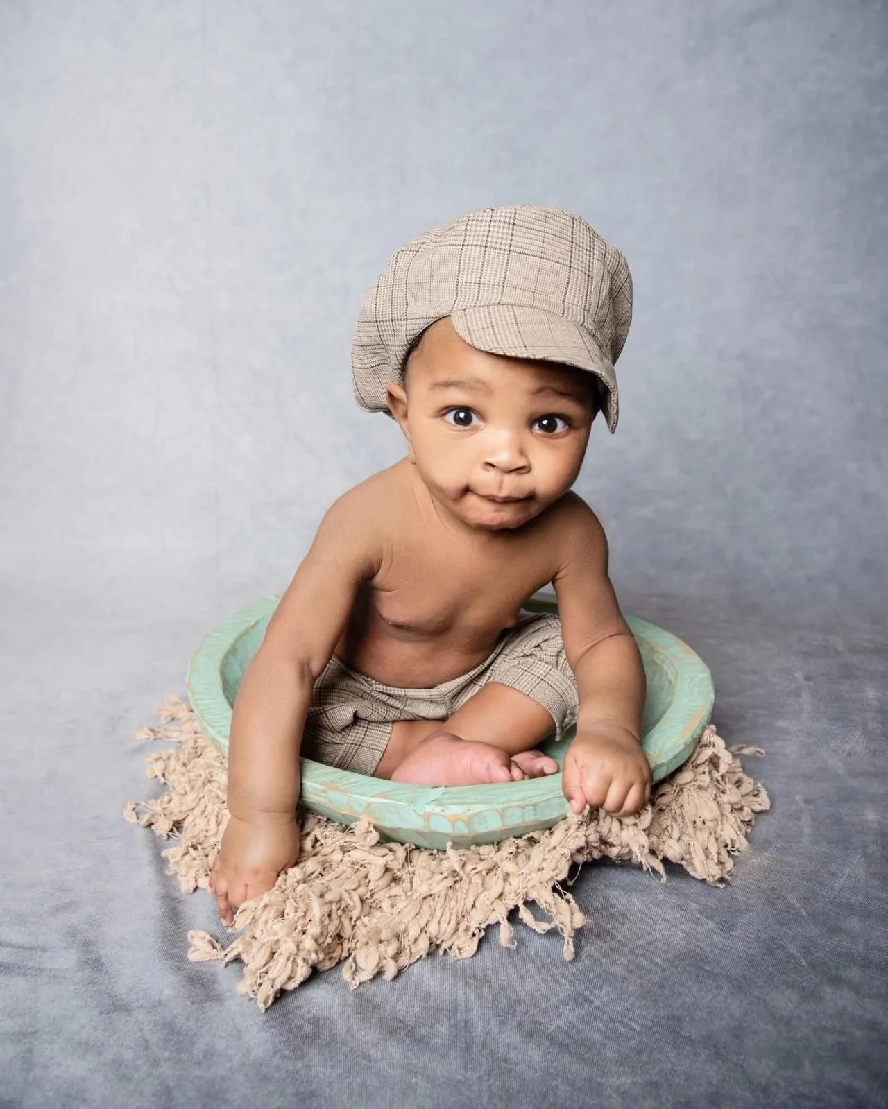 Cute baby boy sitting inside a decorative bowl, wearing a cap and shorts, on a textured mat against a plain gray backdrop.