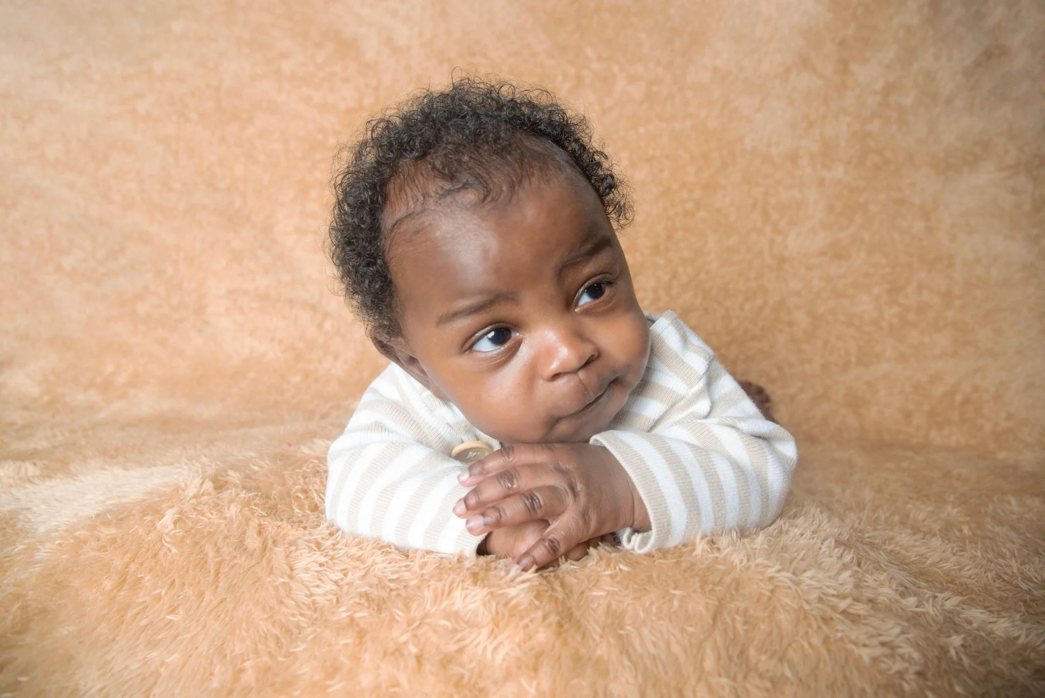 A baby with curly hair lying on a soft beige blanket, resting on his arms and looking to the side.