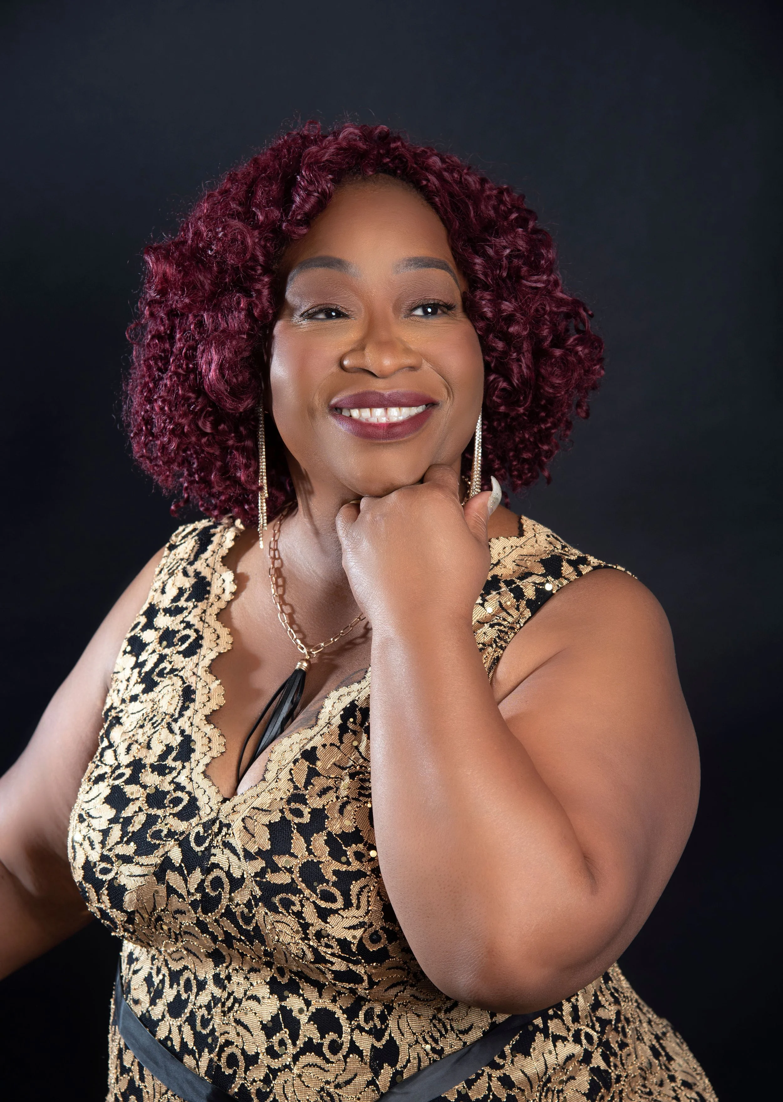 A woman with curly red hair wearing a gold and black leopard print dress and jewelry, smiling with her hand resting under her chin against a black background.