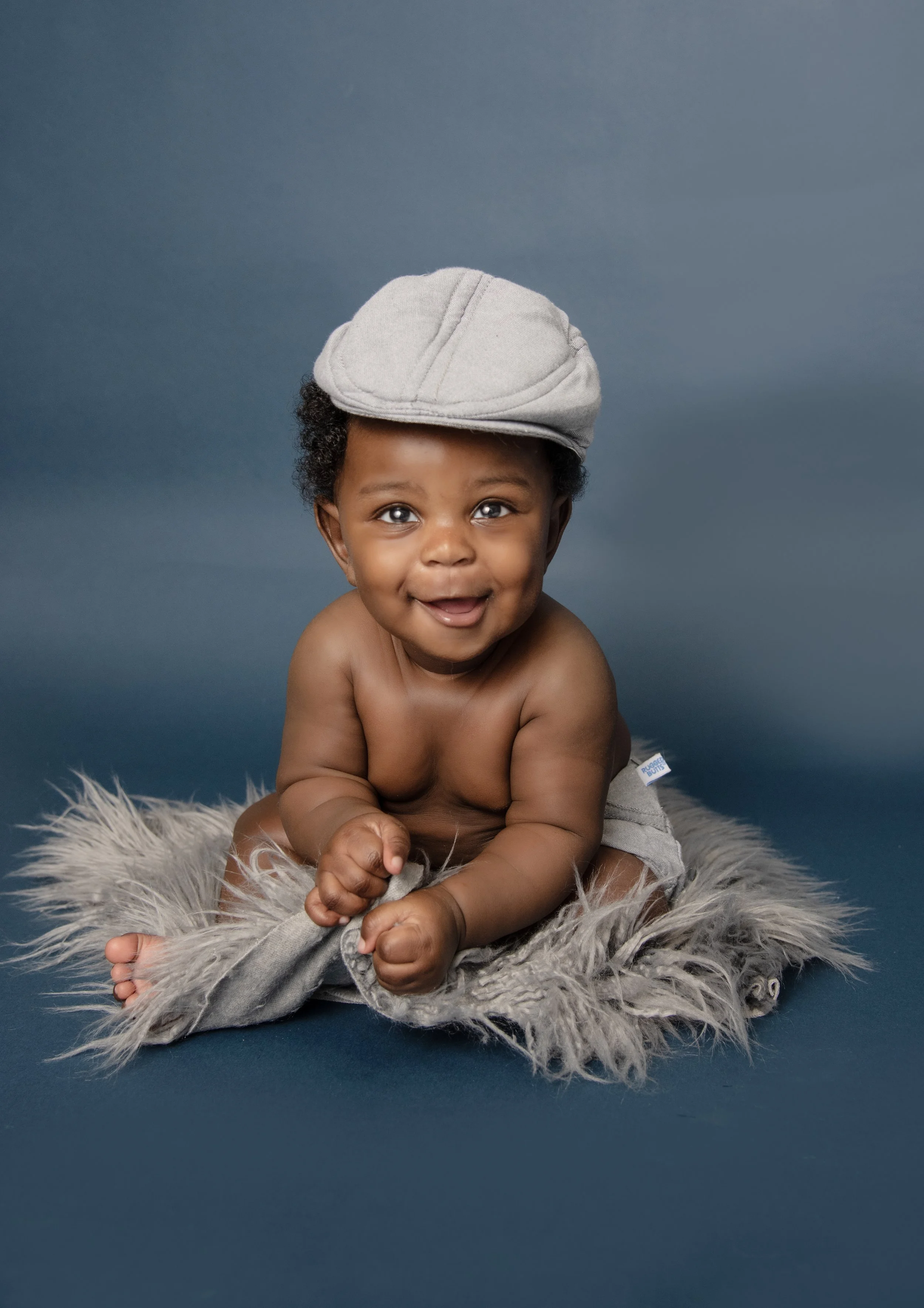 A smiling baby sitting on a fluffy gray rug, wearing a light gray cap, and looking at the camera with a joyful expression.