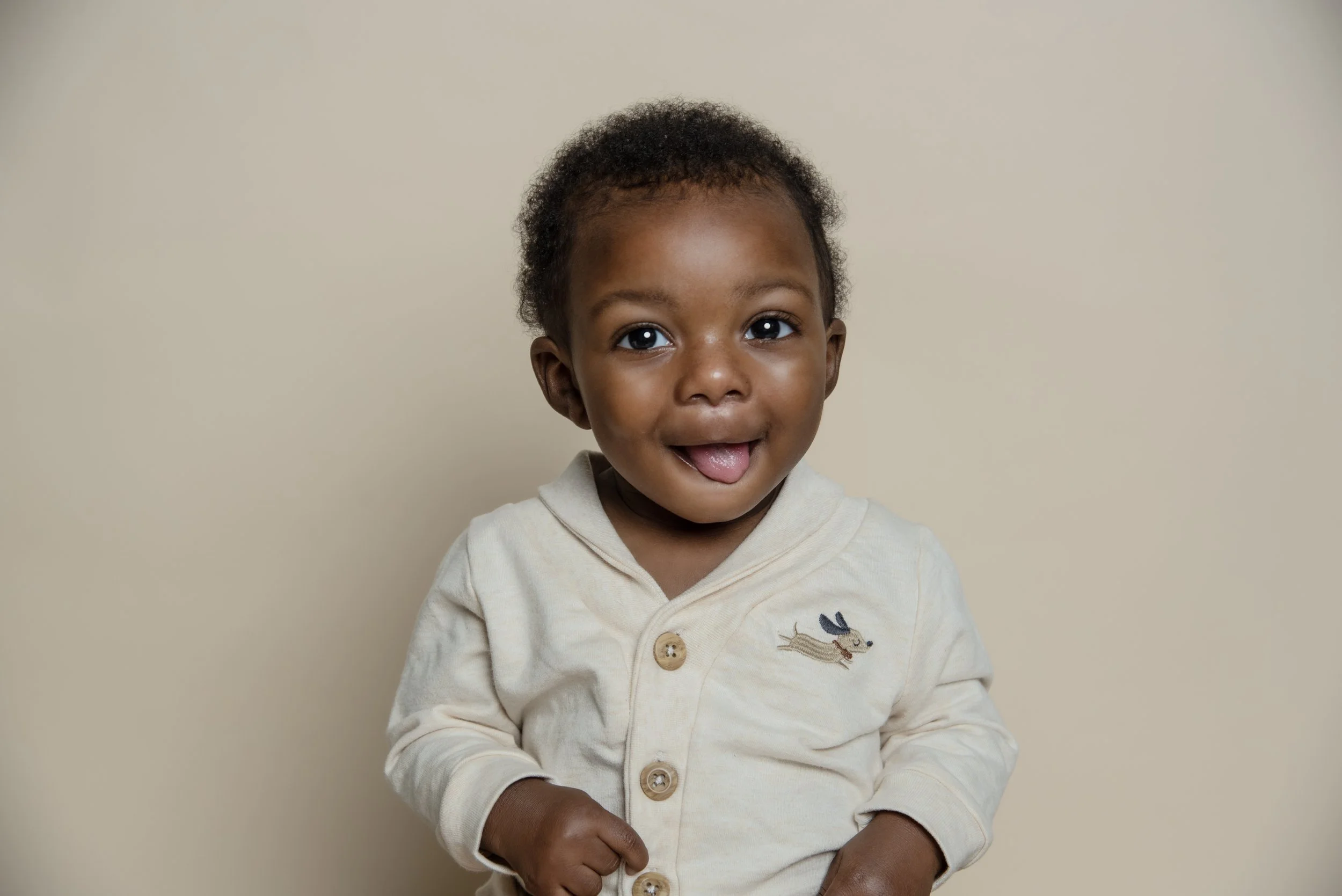 A young African-American child with curly hair smiling and looking at the camera, wearing a light-colored hoodie with a small dog embroidered on it, standing against a plain beige background.