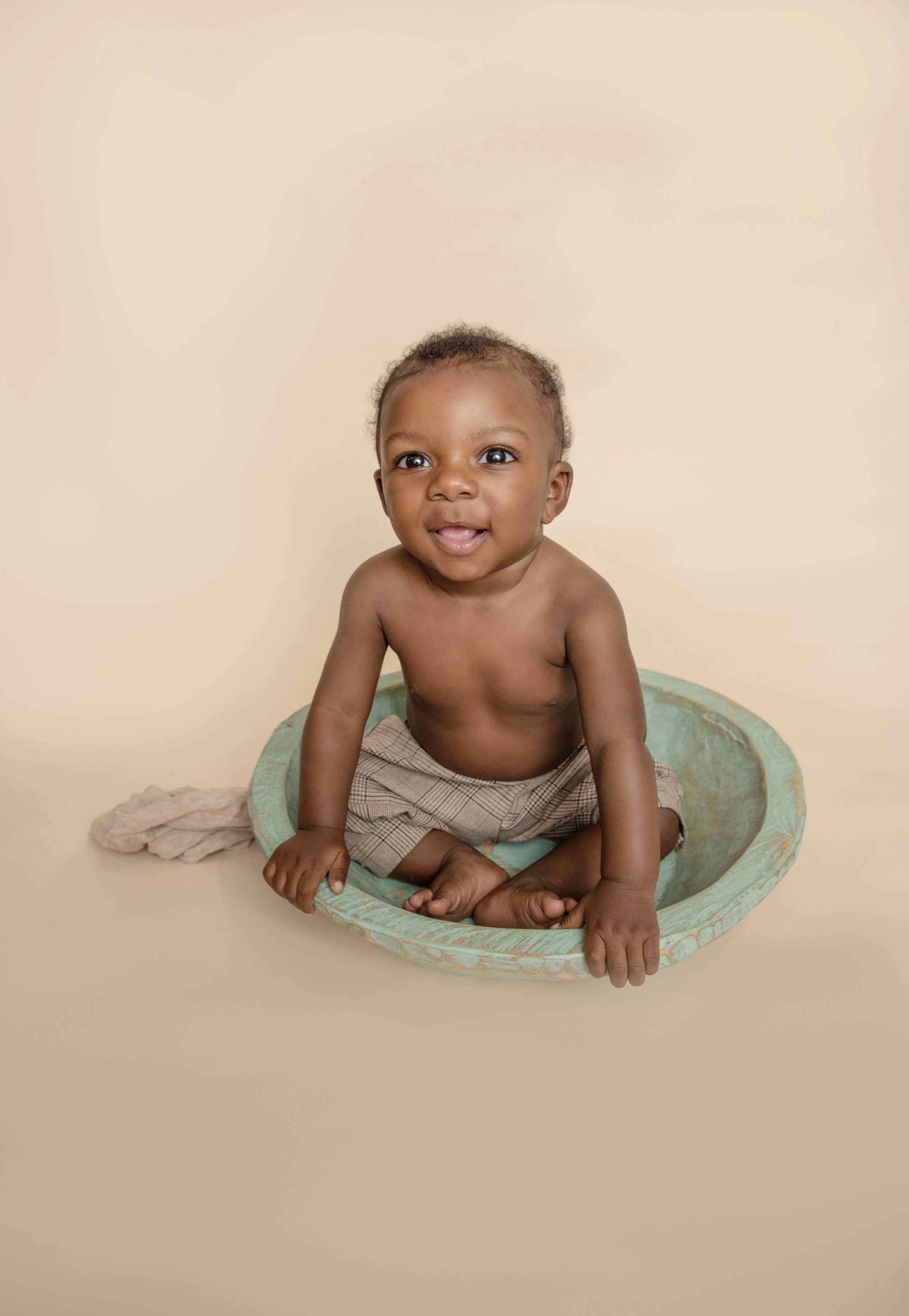 A smiling toddler sitting in a green boat-shaped container on a beige background.