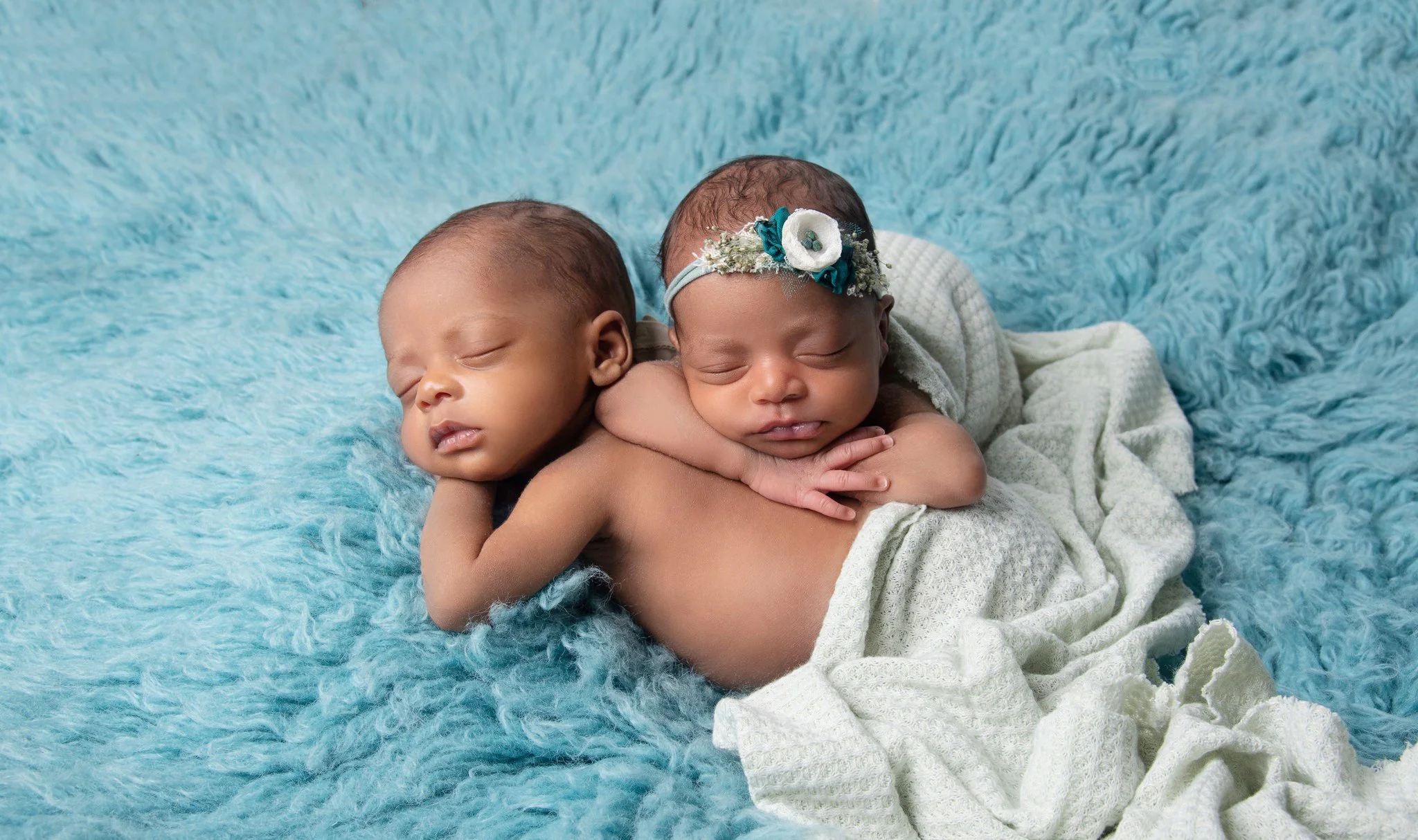Two sleeping infants, one girl and one boy, cuddling on a fluffy blue blanket. The girl is wearing a headband with flowers, and the boy is partially covered with a white knitted blanket.
