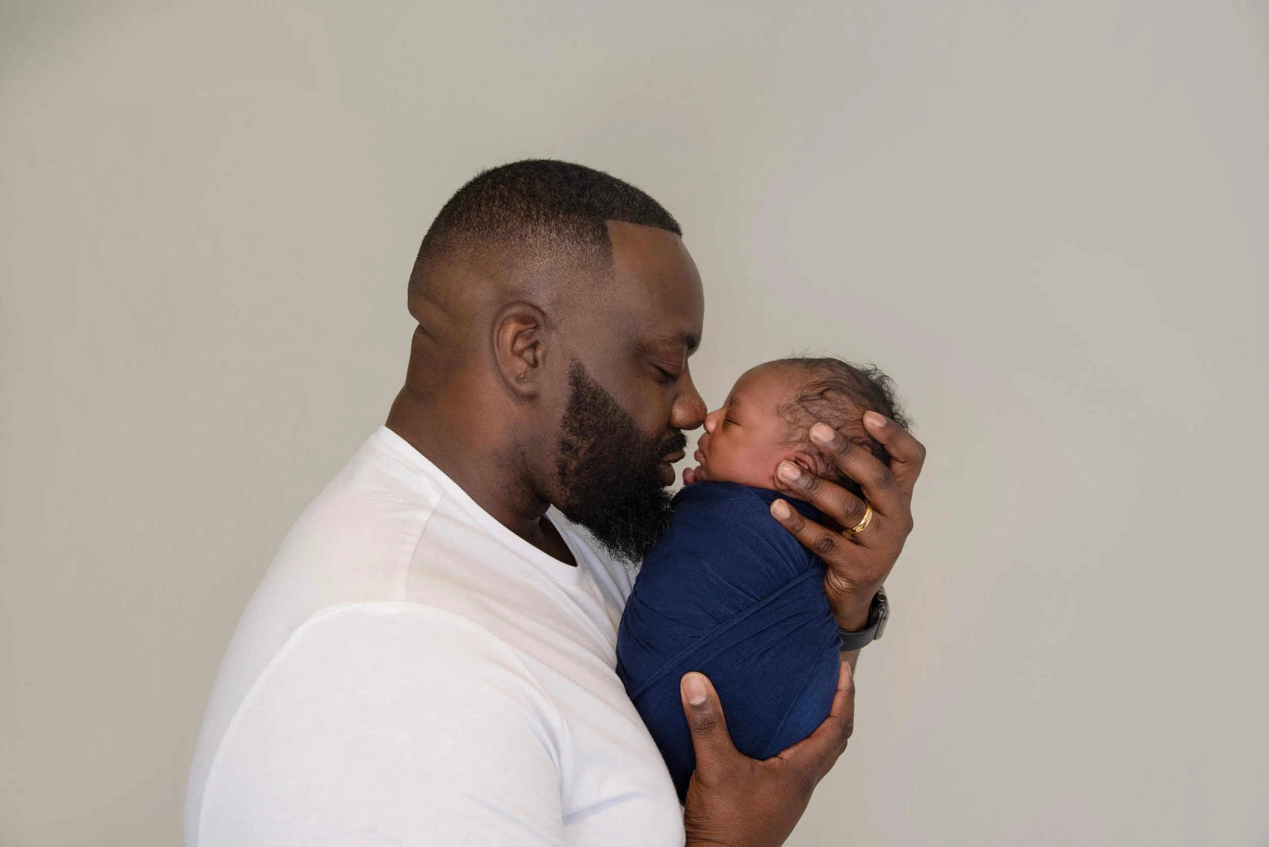 An adult man holding a newborn baby close to his face, gently touching noses, against a plain light-colored background.
