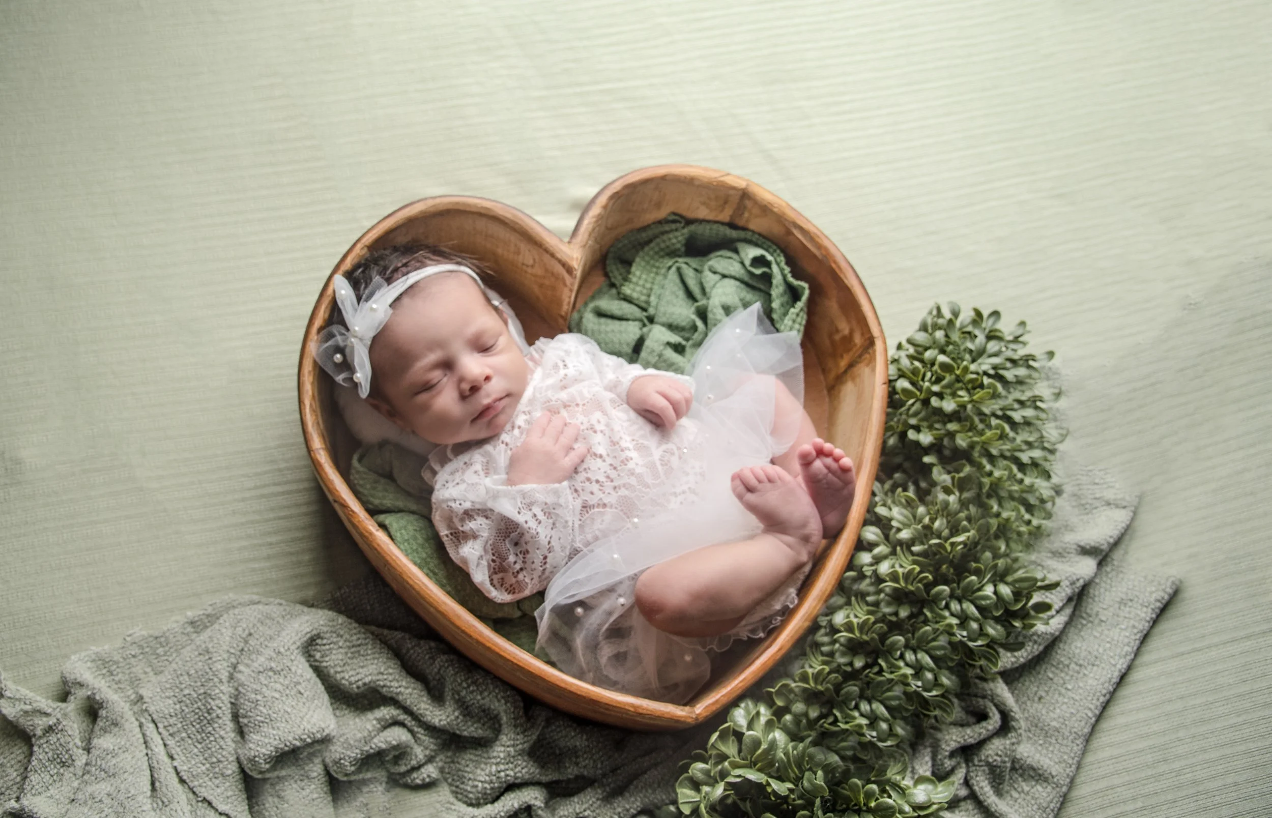Baby girl sleeping in a wooden heart-shaped basket with green blanket and artificial plants nearby