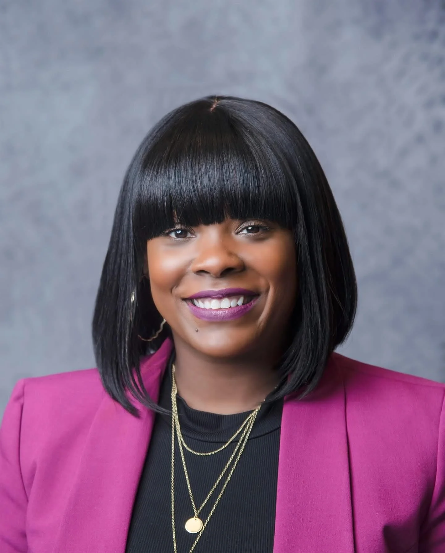 A portrait of a woman with straight black hair, wearing a pink blazer and black top, smiling, with layered gold necklaces and purple lipstick, against a gray background for school headshot.