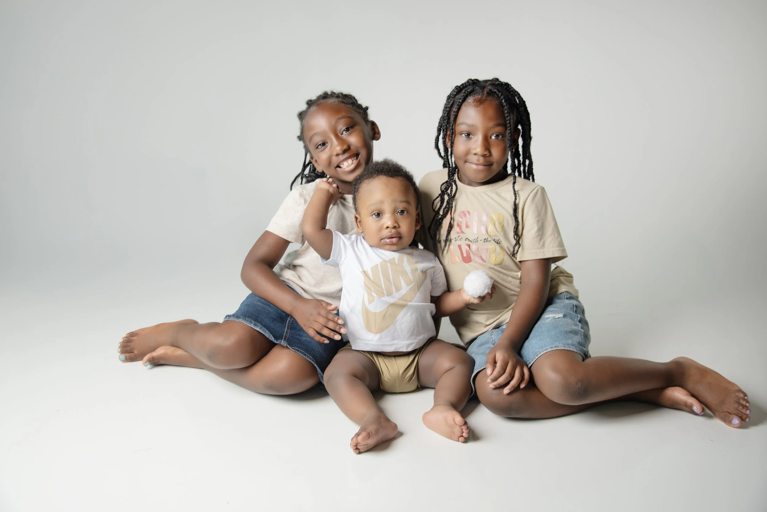 Three children sitting on the floor against a plain background, smiling and looking at the camera.