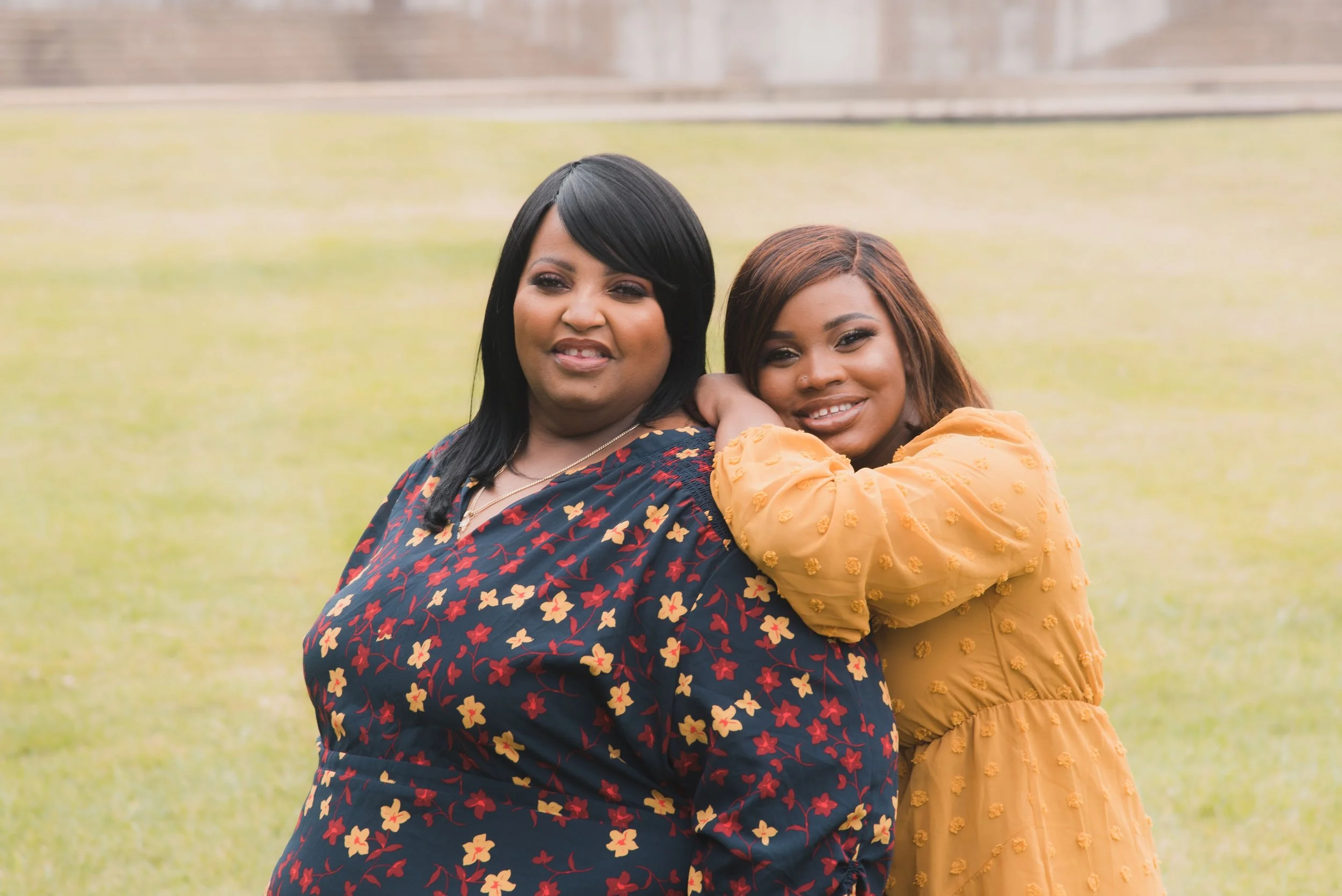 Two women standing outside on grass, smiling. One woman has black hair and wears a navy dress with red and yellow floral pattern, the other has light brown hair and wears a yellow dress with textured details, with her arm resting on the first woman's