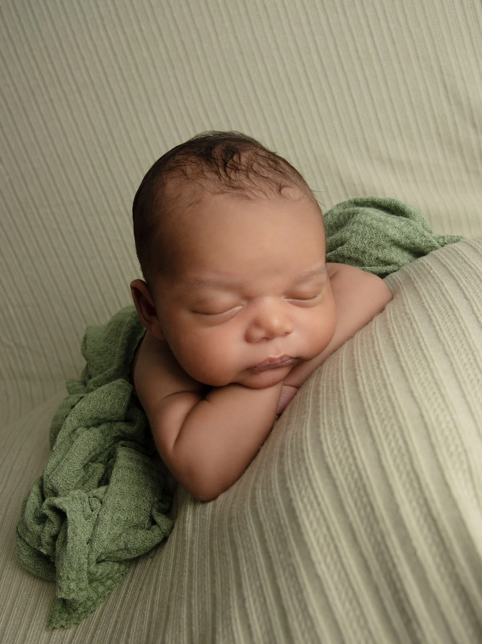 A sleeping infant with his head resting on his arms, lying on a striped beige pillow, with a green cloth nearby and a green textured blanket draped over his back.