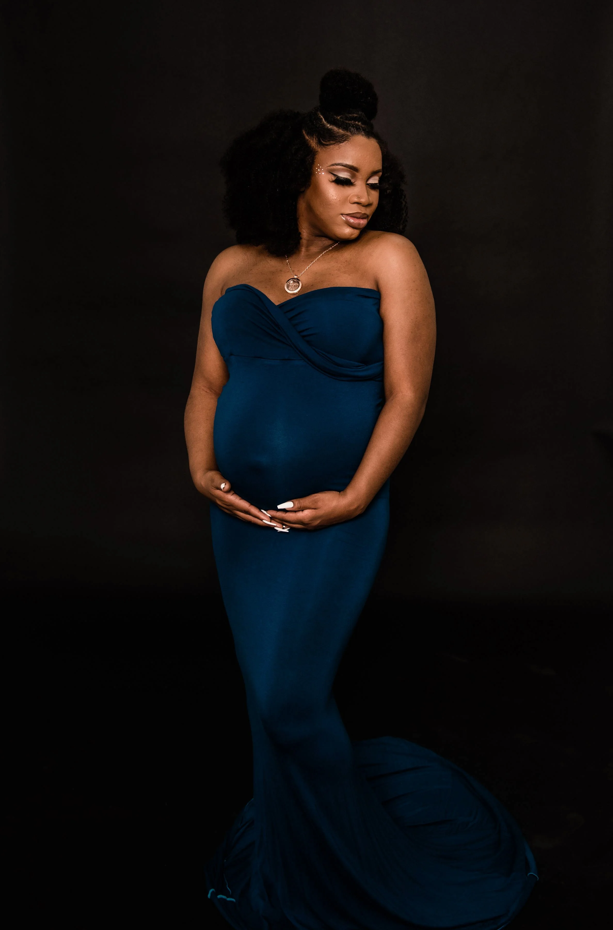 A pregnant woman in a strapless blue dress posed against a black background, holding her belly with both hands, with natural hair styled in a curly afro.
