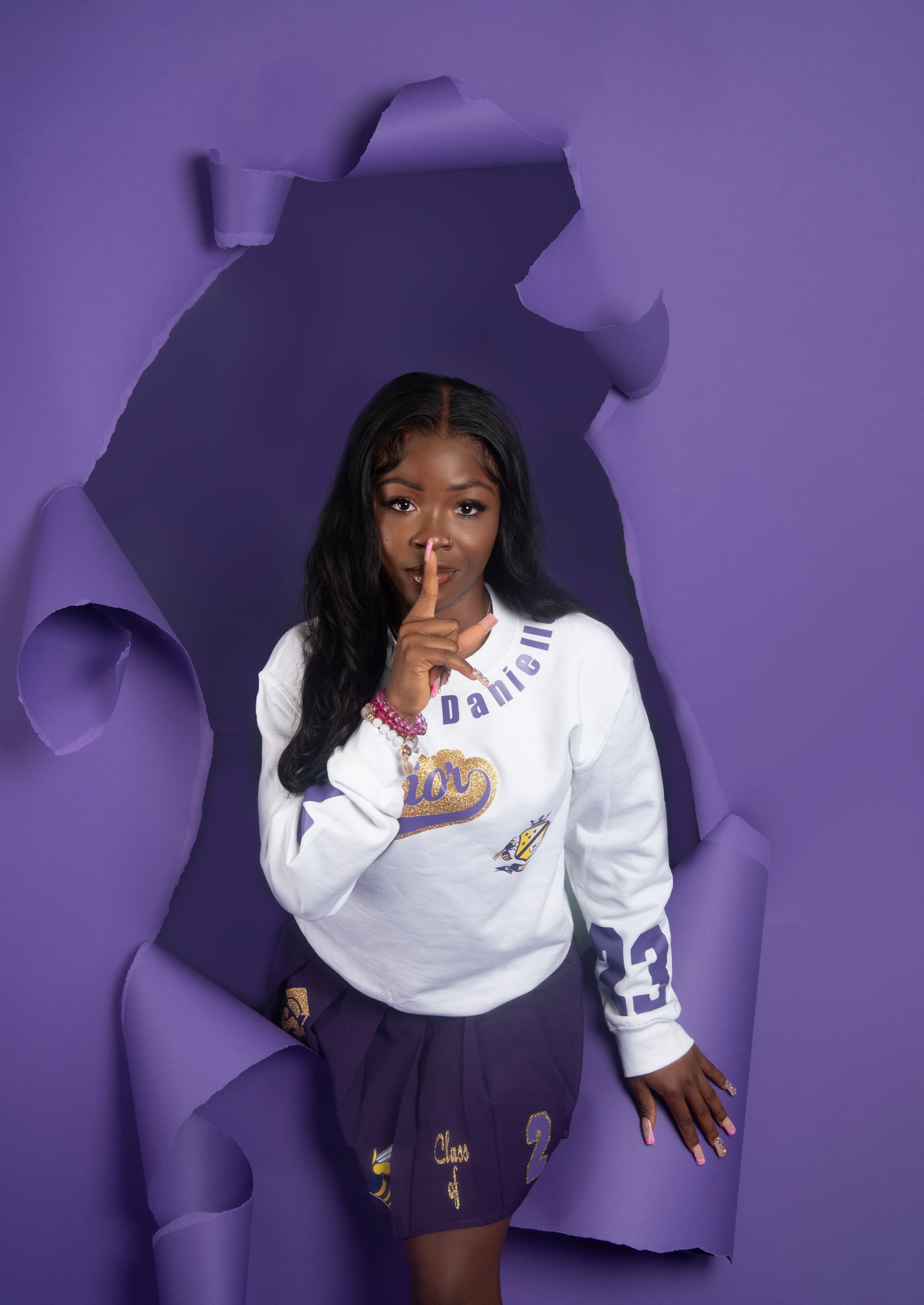 A woman standing in front of a purple paper background with a torn hole, holding her finger up to her lips. She is wearing a white sweatshirt with purple and gold accents, including the word "Dance" and the number "23," as well as a purple skirt with