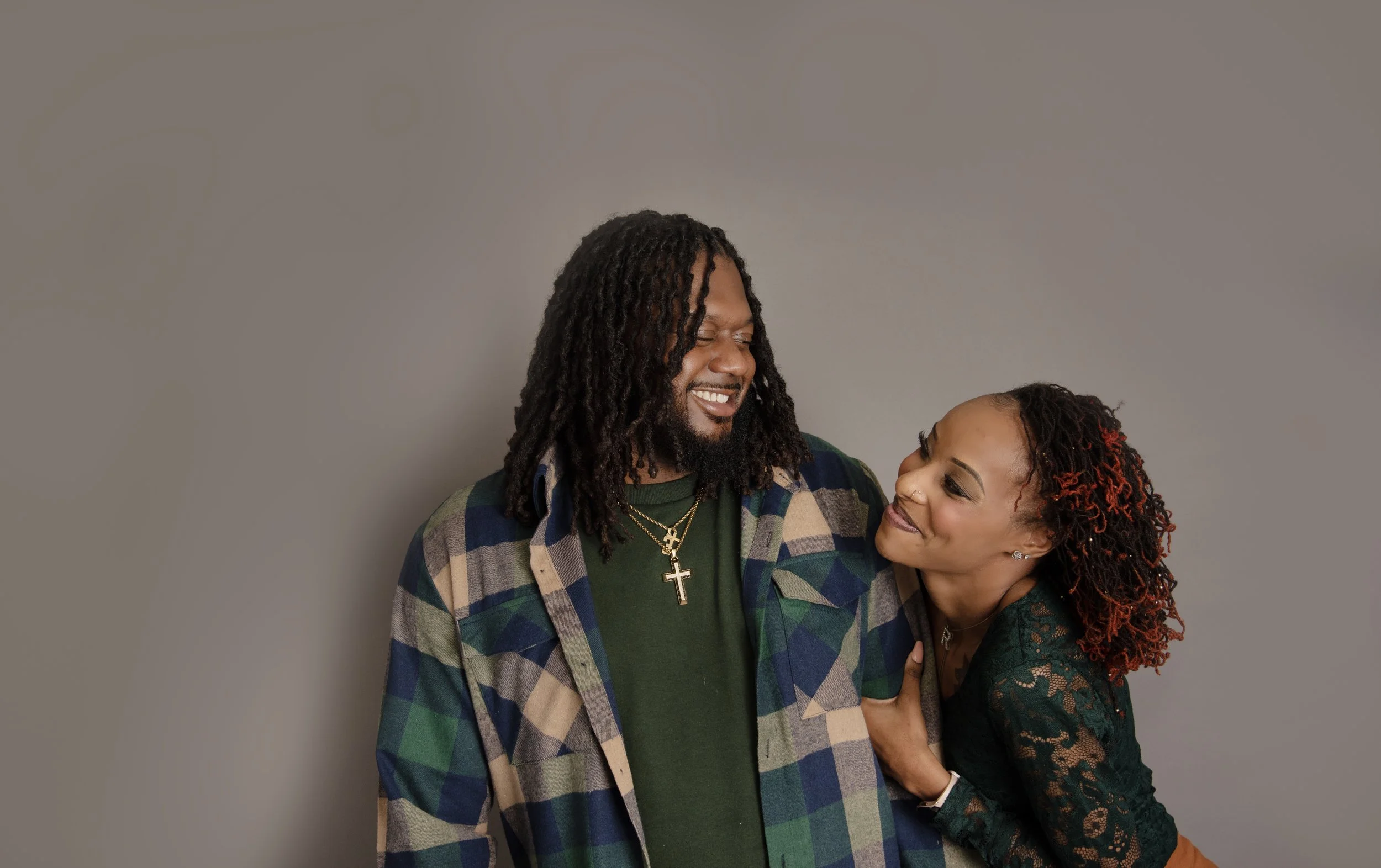 A smiling man with dreadlocks and a woman with curly hair and red highlights, both looking at each other affectionately against a plain gray background.