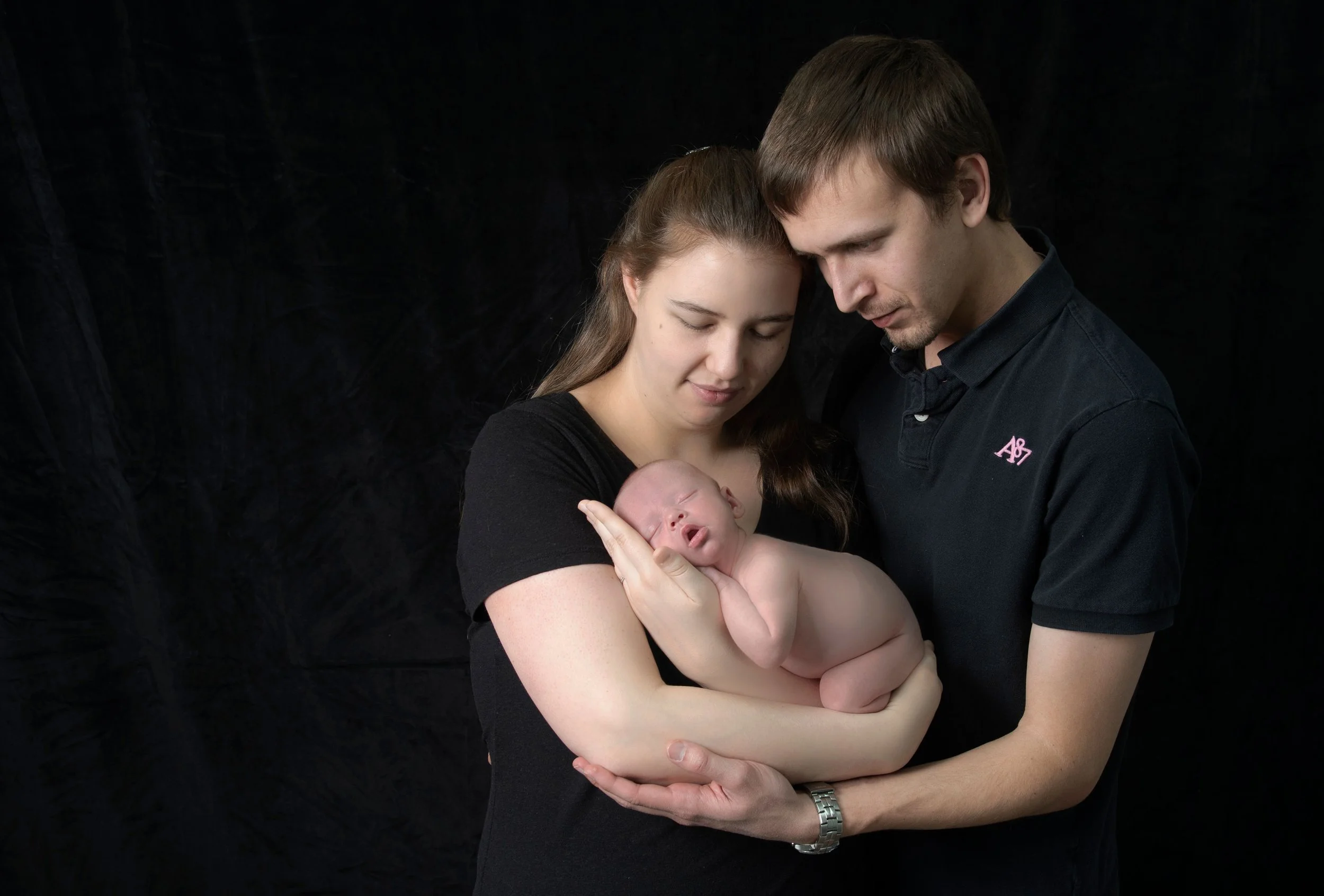 A young couple holding a newborn baby against a black background.