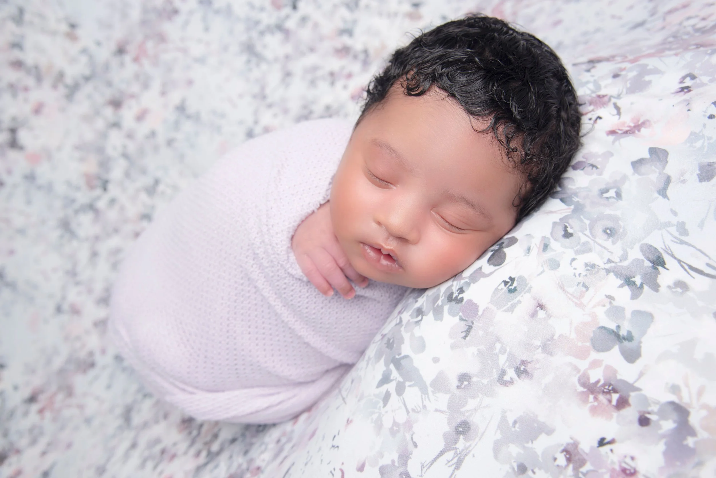 A sleeping baby with dark curly hair resting on a floral pillow and grey speckled blanket.
