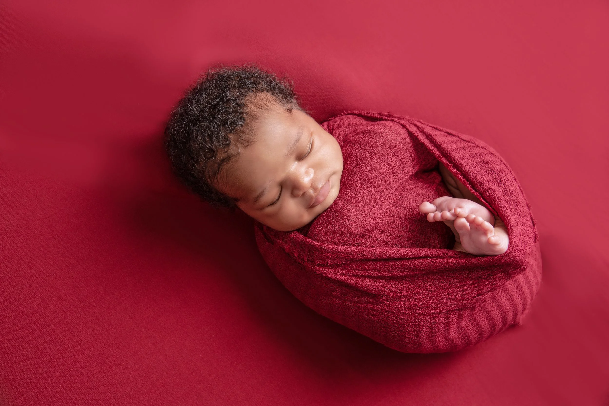 A sleeping baby swaddled in a red blanket on a matching red background.