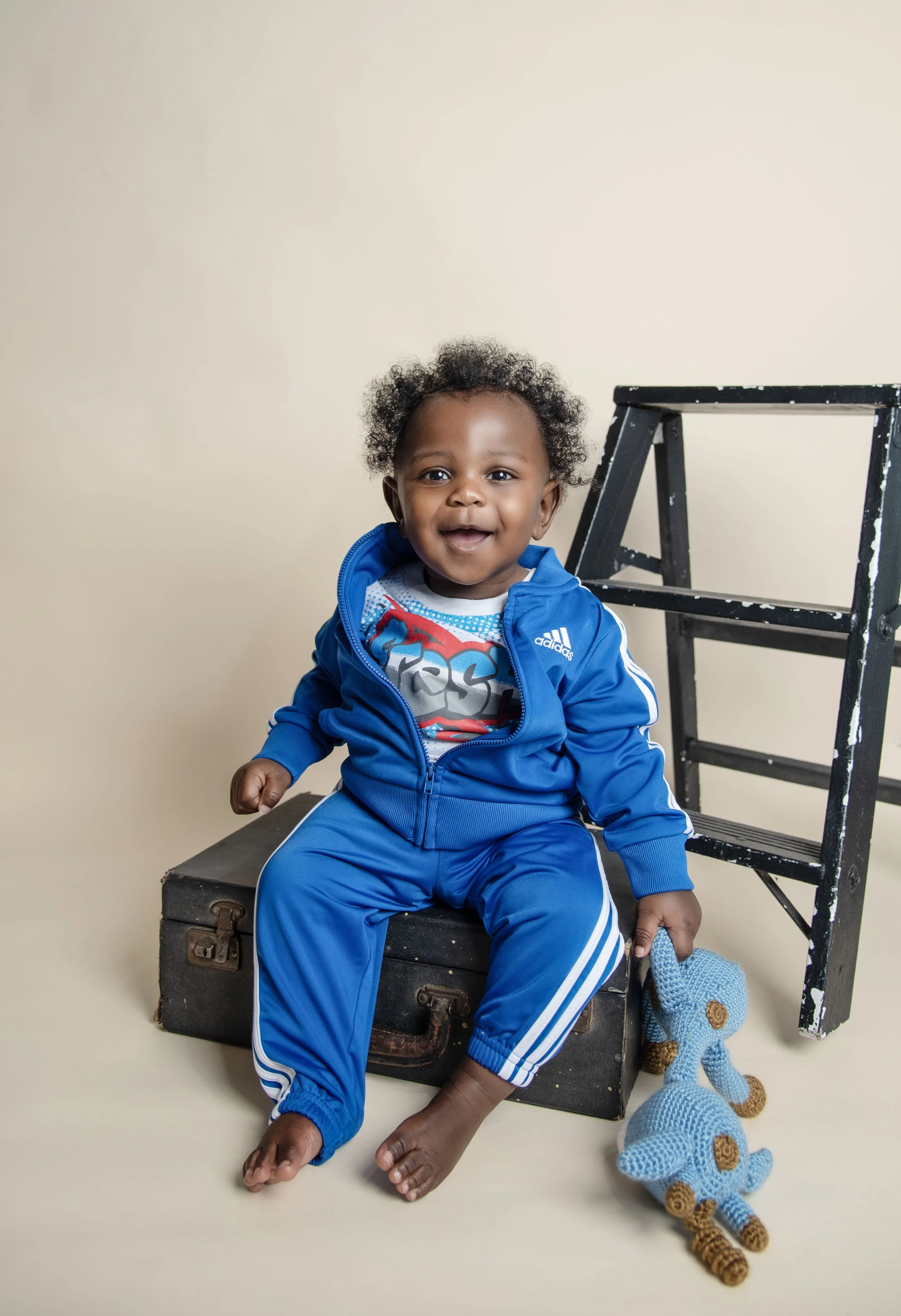 A joyful young child with curly hair dressed in a blue athletic outfit sitting on a vintage suitcase, holding a crocheted blue teddy bear, with a step stool behind him against a plain beige background.