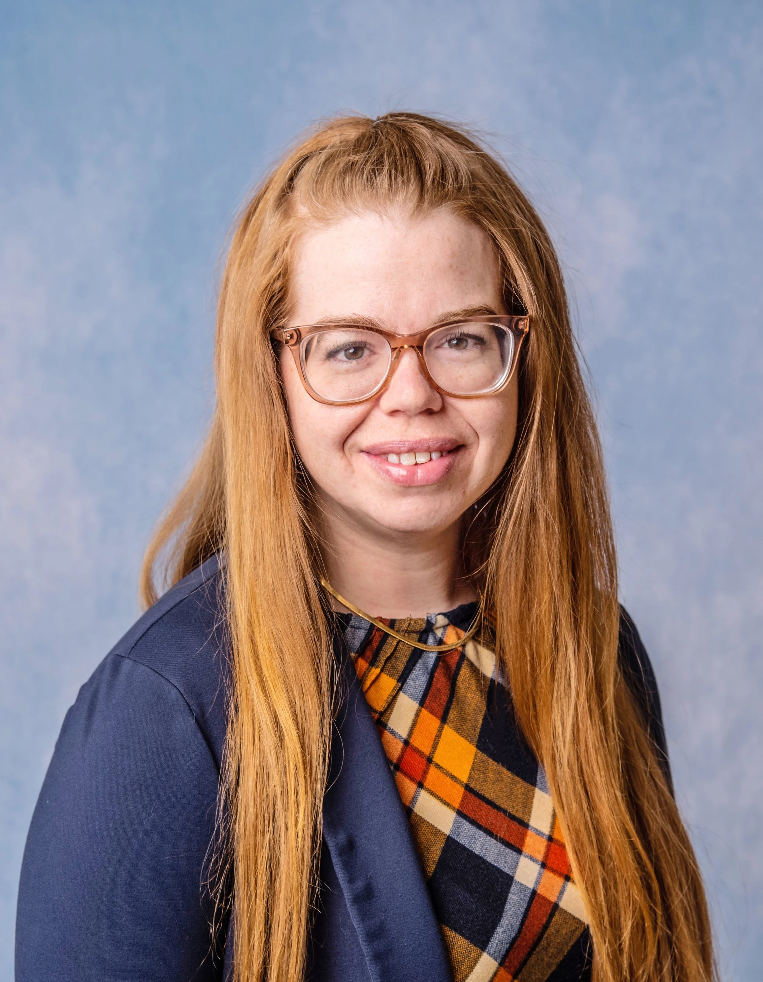 Head-and-shoulders portrait of a woman with long red hair, wearing glasses, a plaid shirt, and a dark blazer, against a blue background.