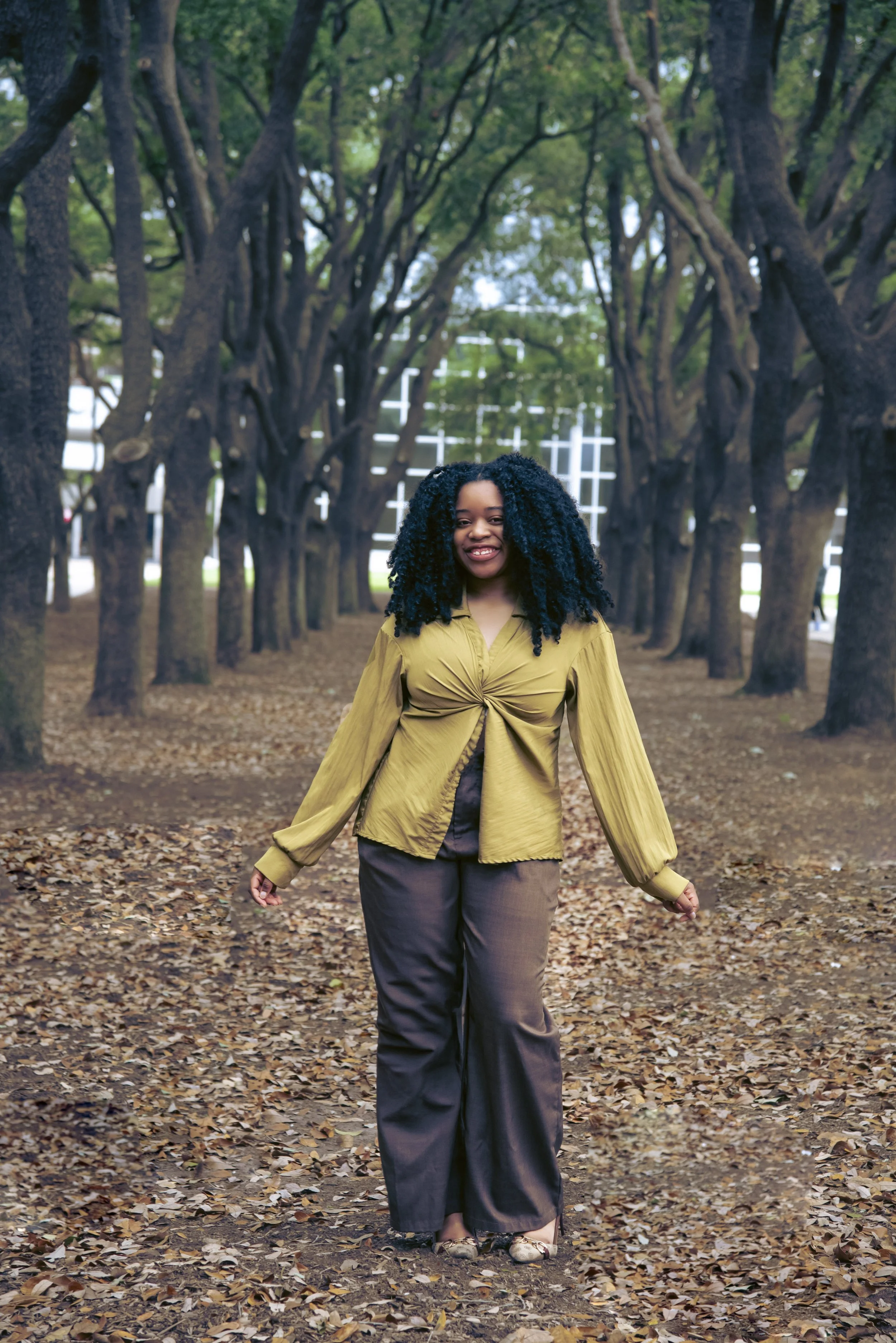 A woman with curly black hair smiling while walking on a leaf-covered path through a park with tall trees and green foliage.