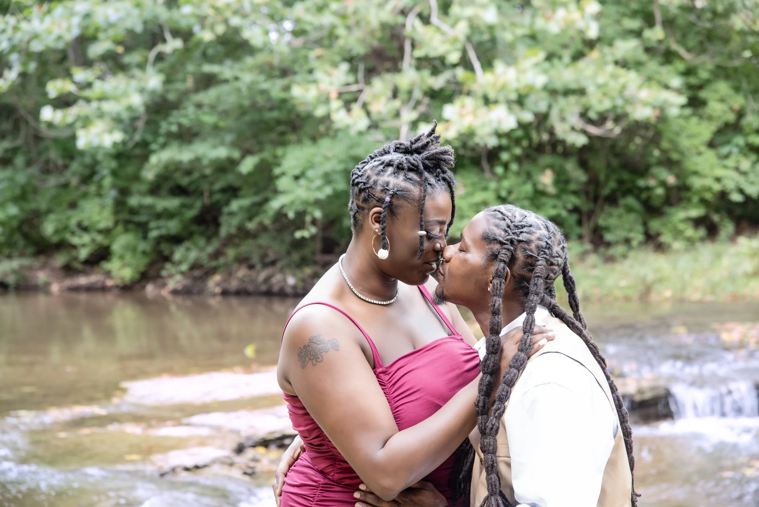 A couple with dreadlocks kissing outdoors near a creek, surrounded by green trees.