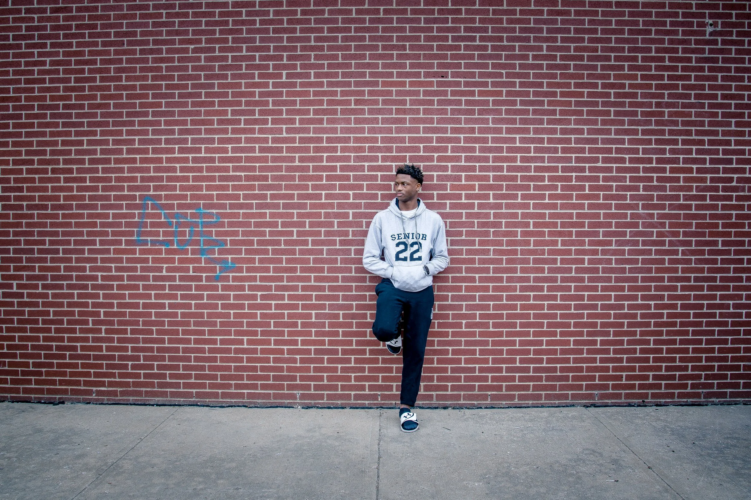 A young man in a gray hoodie with the words 'Senior 22' stands against a red brick wall, with one foot resting on the wall and hands in pockets.