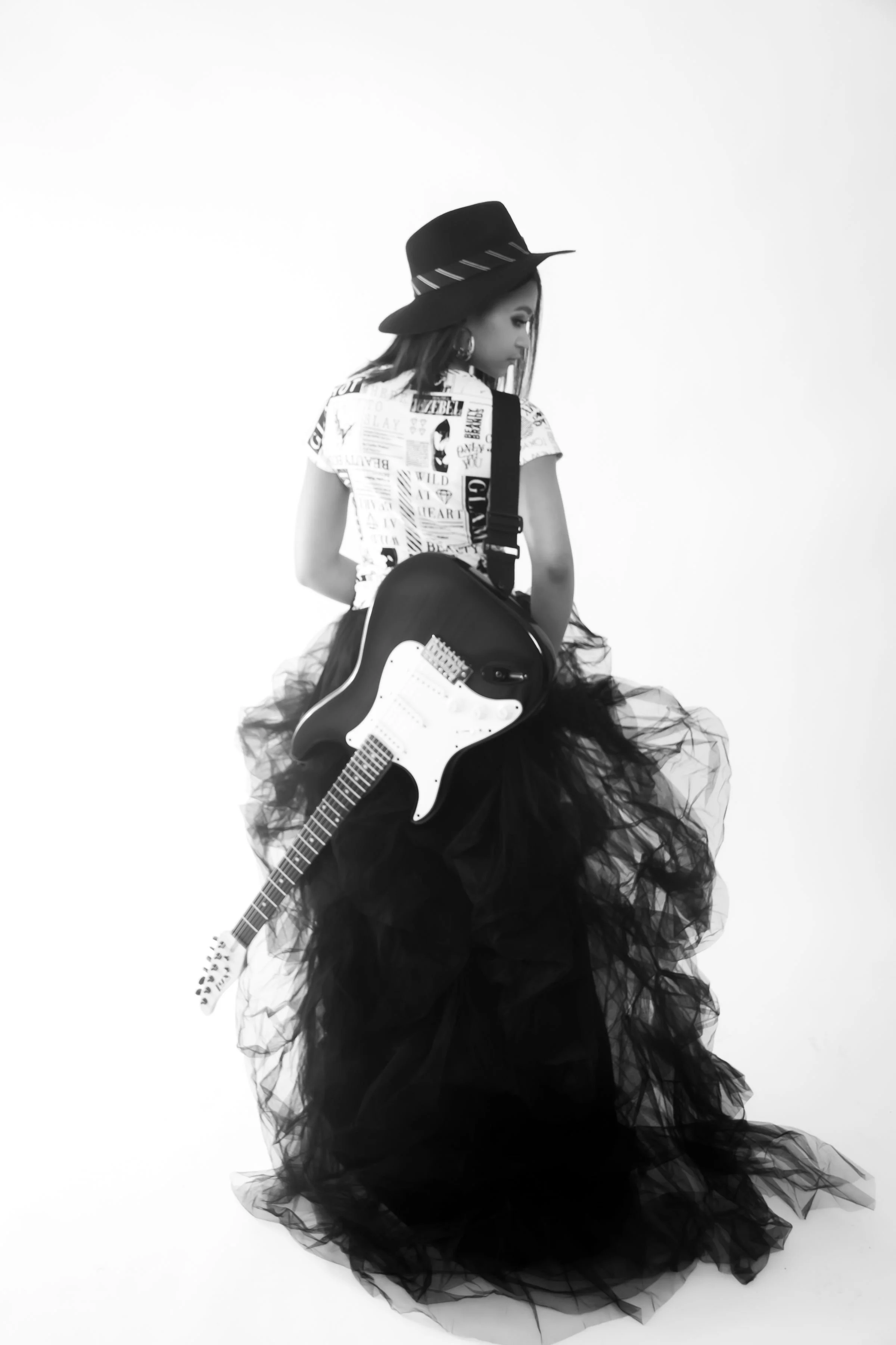 A woman wearing a wide-brimmed hat, t-shirt with newspaper print, and a voluminous black tulle skirt is holding an electric guitar.