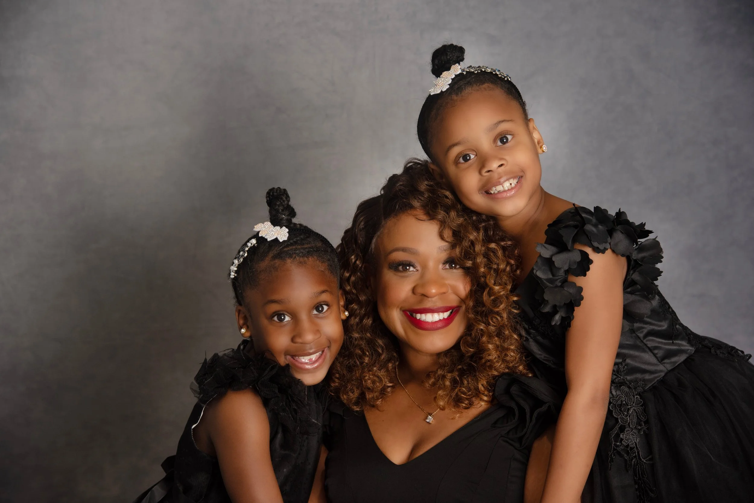 A woman with curly hair and red lipstick smiling, flanked by two young girls, all dressed in black with flower details, posing against a gray background.