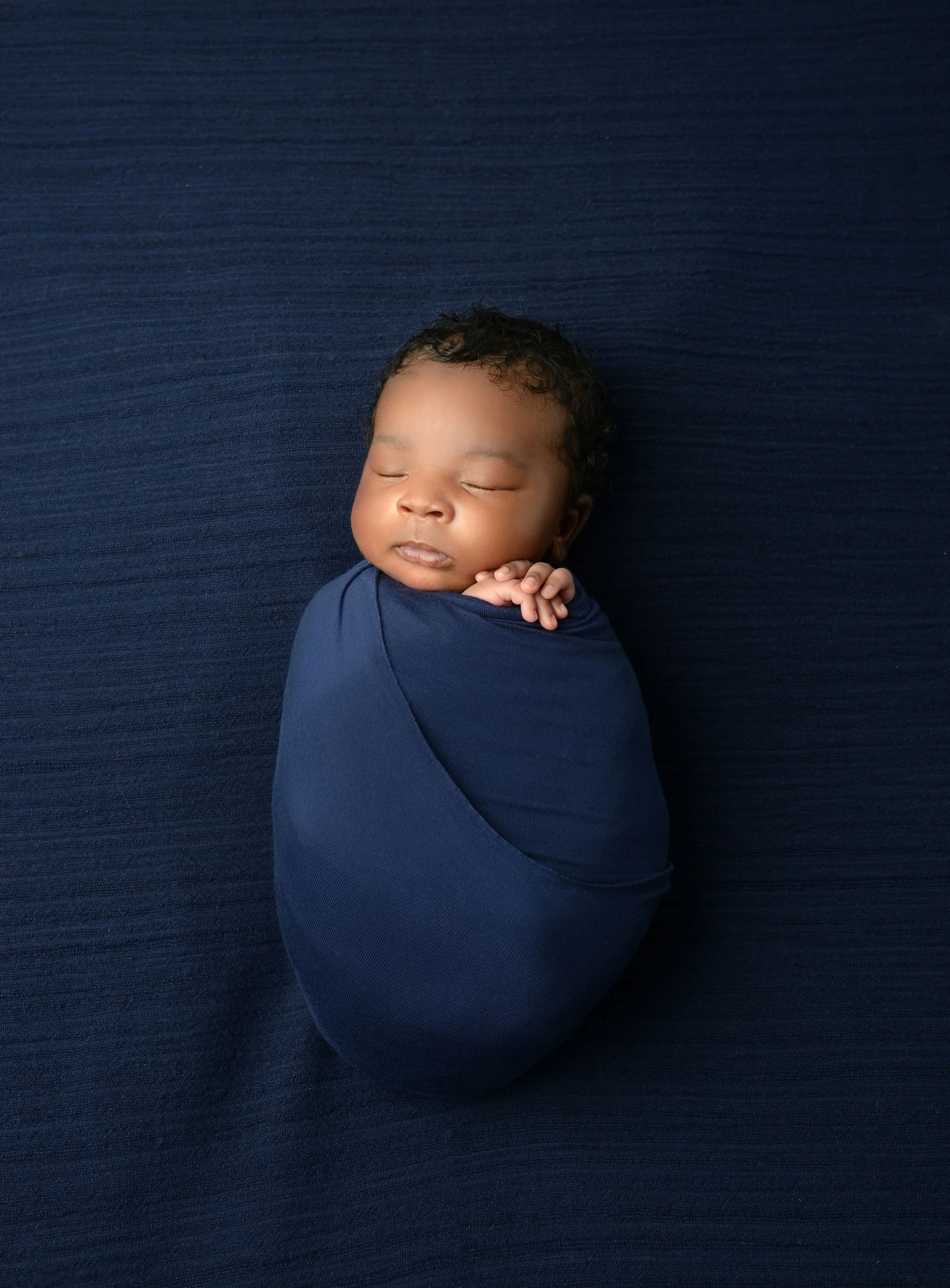 A sleeping baby wrapped in a navy blue blanket, lying on a matching navy blue surface.