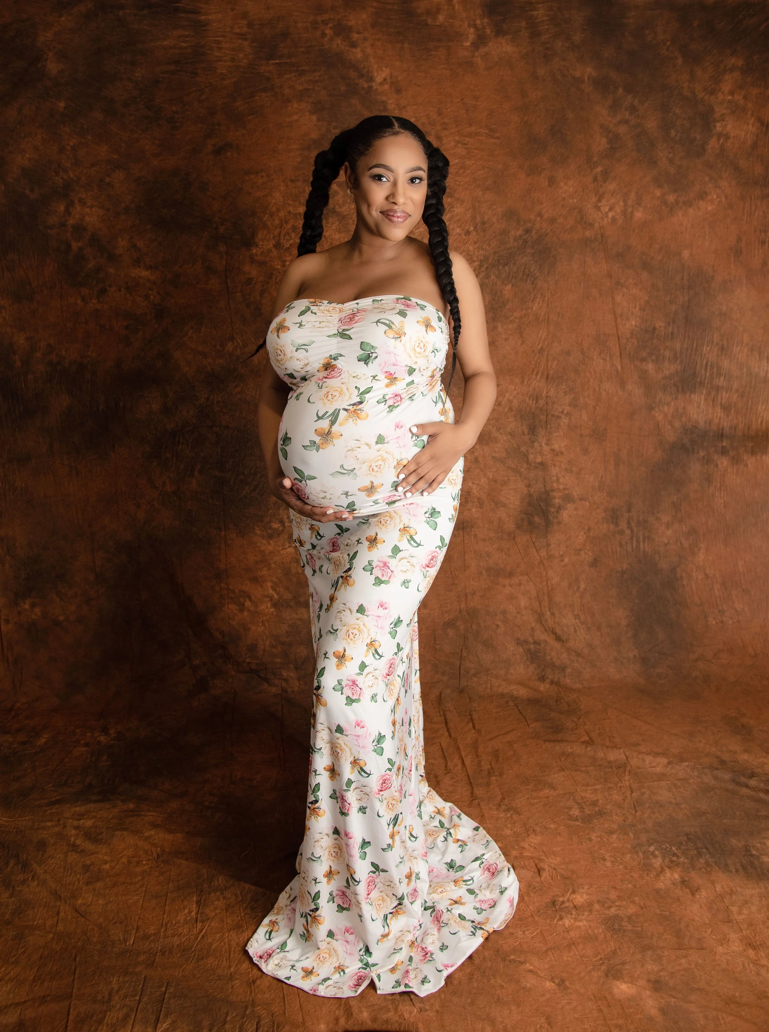 Pregnant woman with long braided hair in a floral strapless gown standing against a brown textured background.