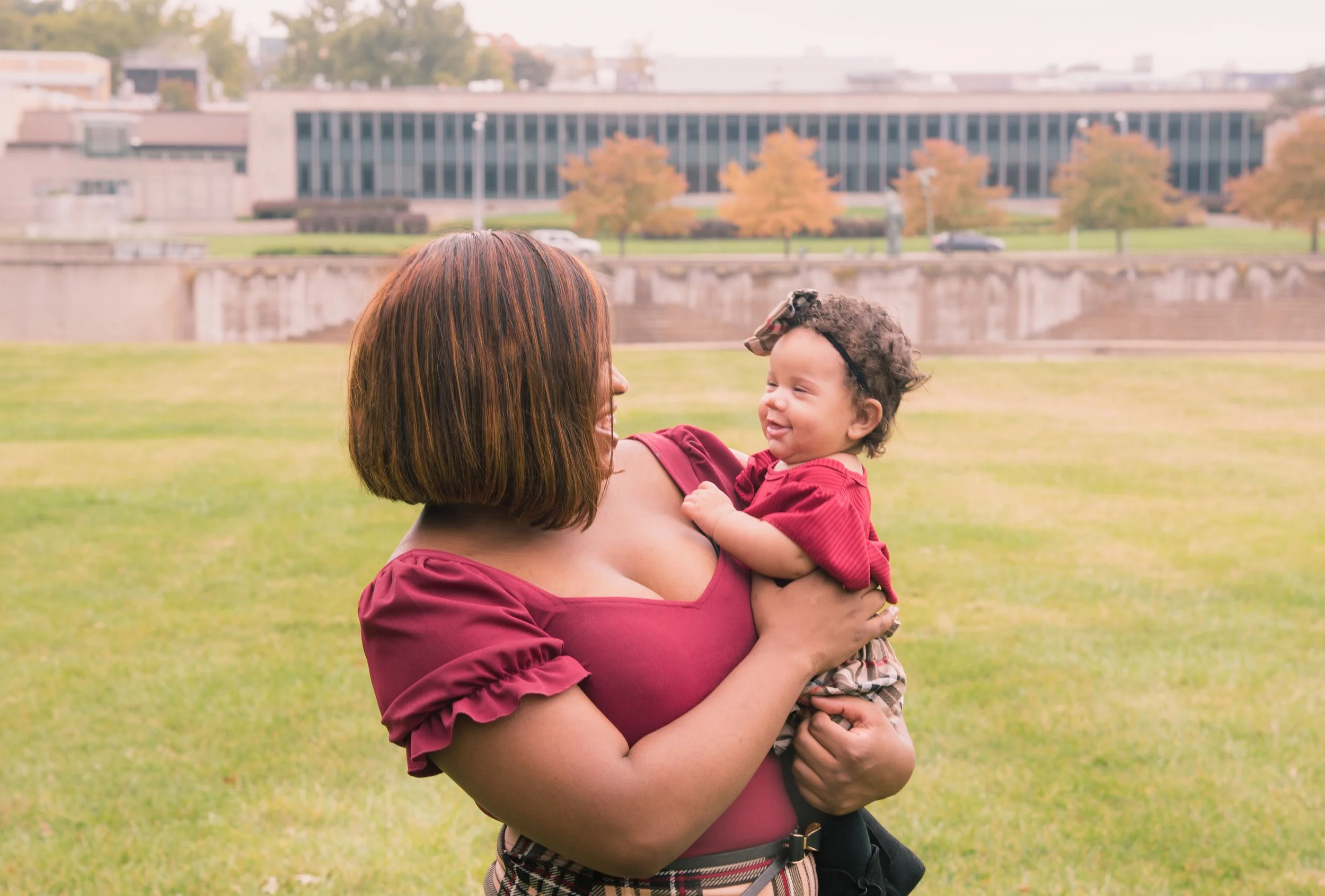 A woman holding a smiling toddler girl outdoors on a grassy field with buildings and trees with fall foliage in the background.