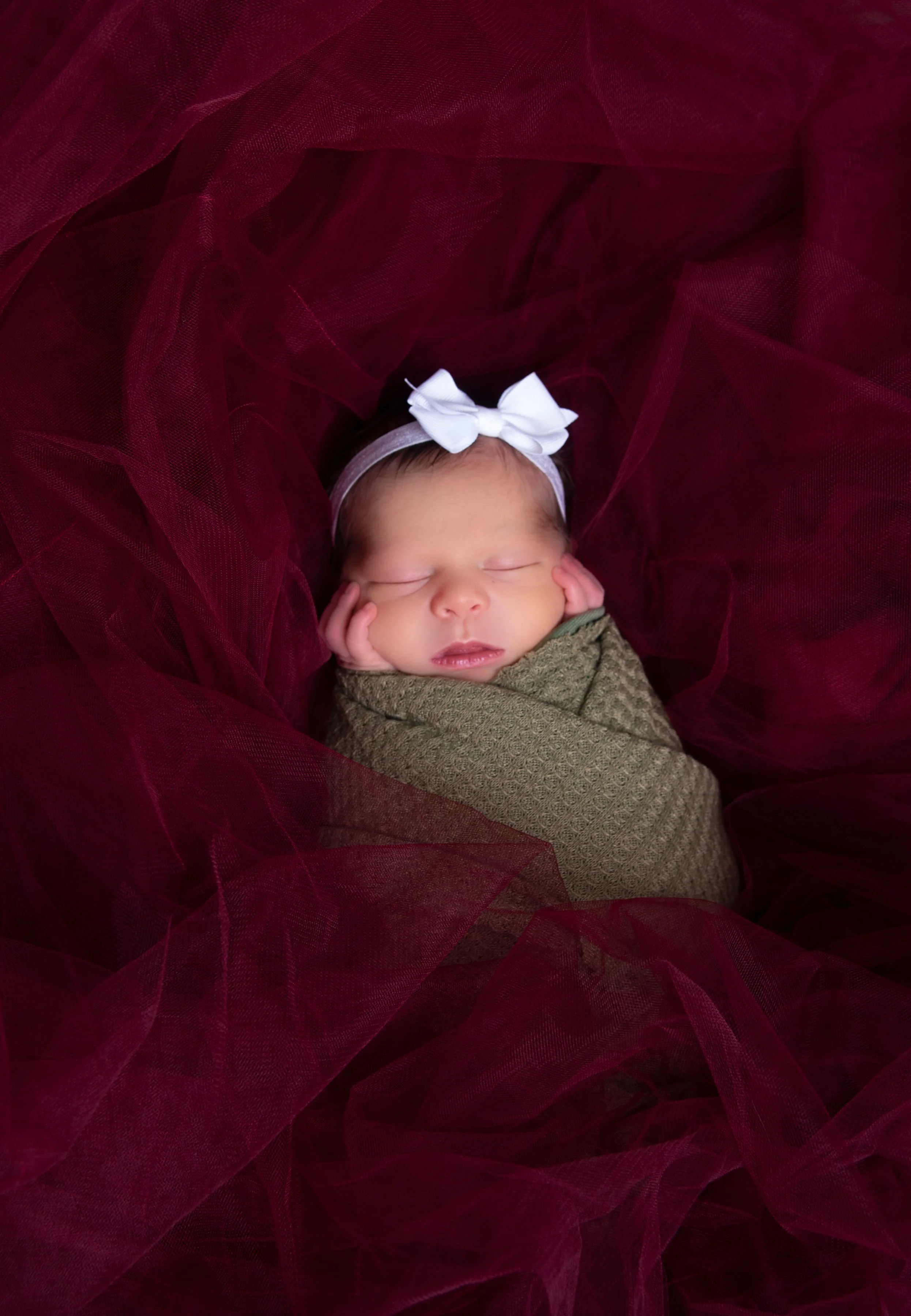 A sleeping baby wrapped in a green blanket, wearing a white bow headband, surrounded by burgundy tulle fabric.