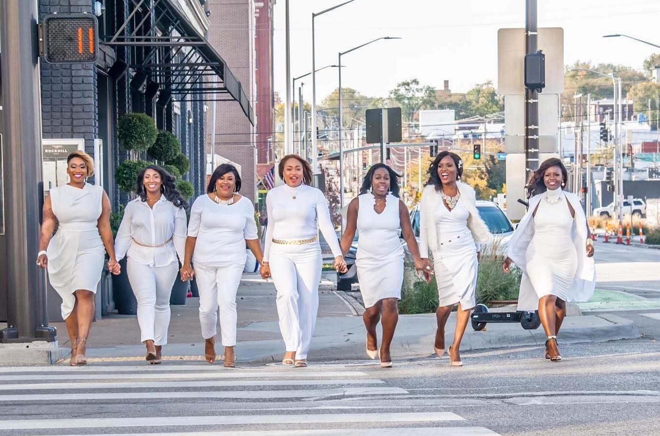 Group of hairstylists dressed in white walking across a city crosswalk, smiling and holding hands, with cars and streetlights in the background for a branding session
